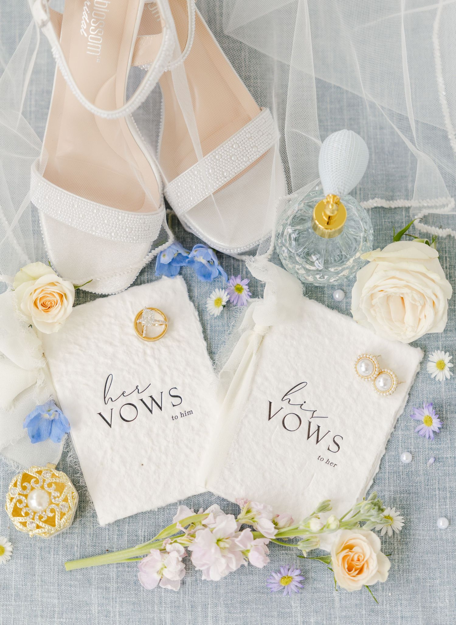 Elegant wedding flat lay with white heels, vow books, rings, earrings, perfume, and blue and white flowers.