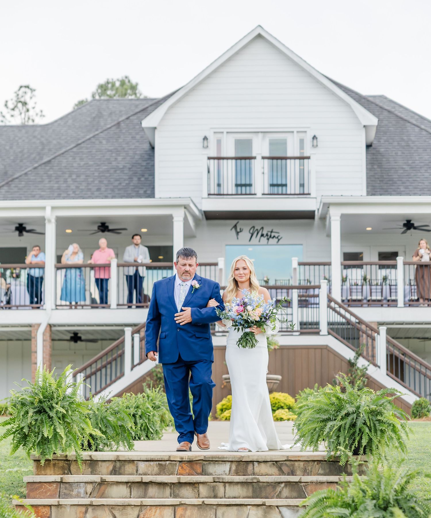 Father in navy suit walks bride holding colorful bouquet down steps at white farmhouse wedding venue.
