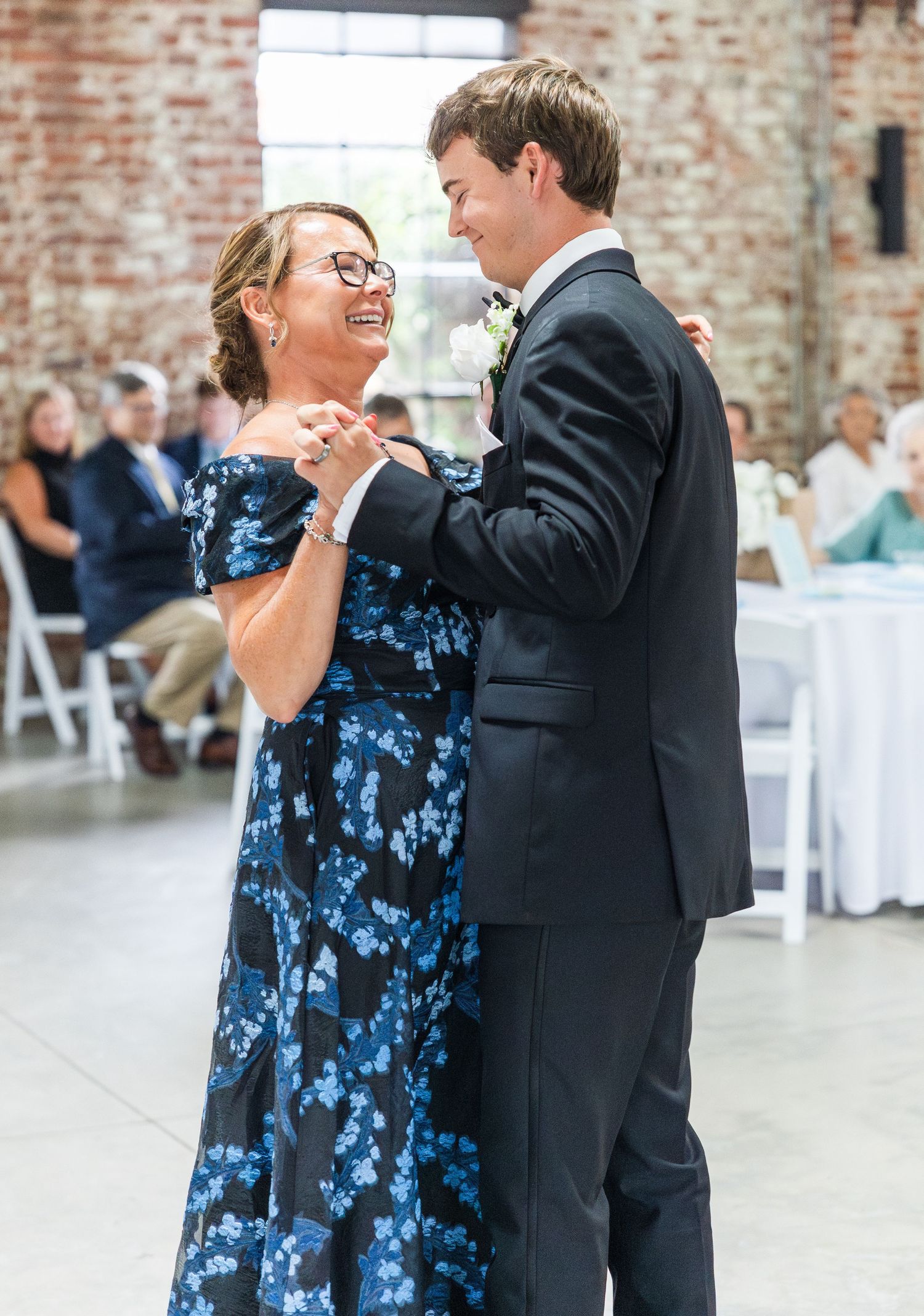Groom dances with mother in blue floral dress during mother-son dance at wedding reception.