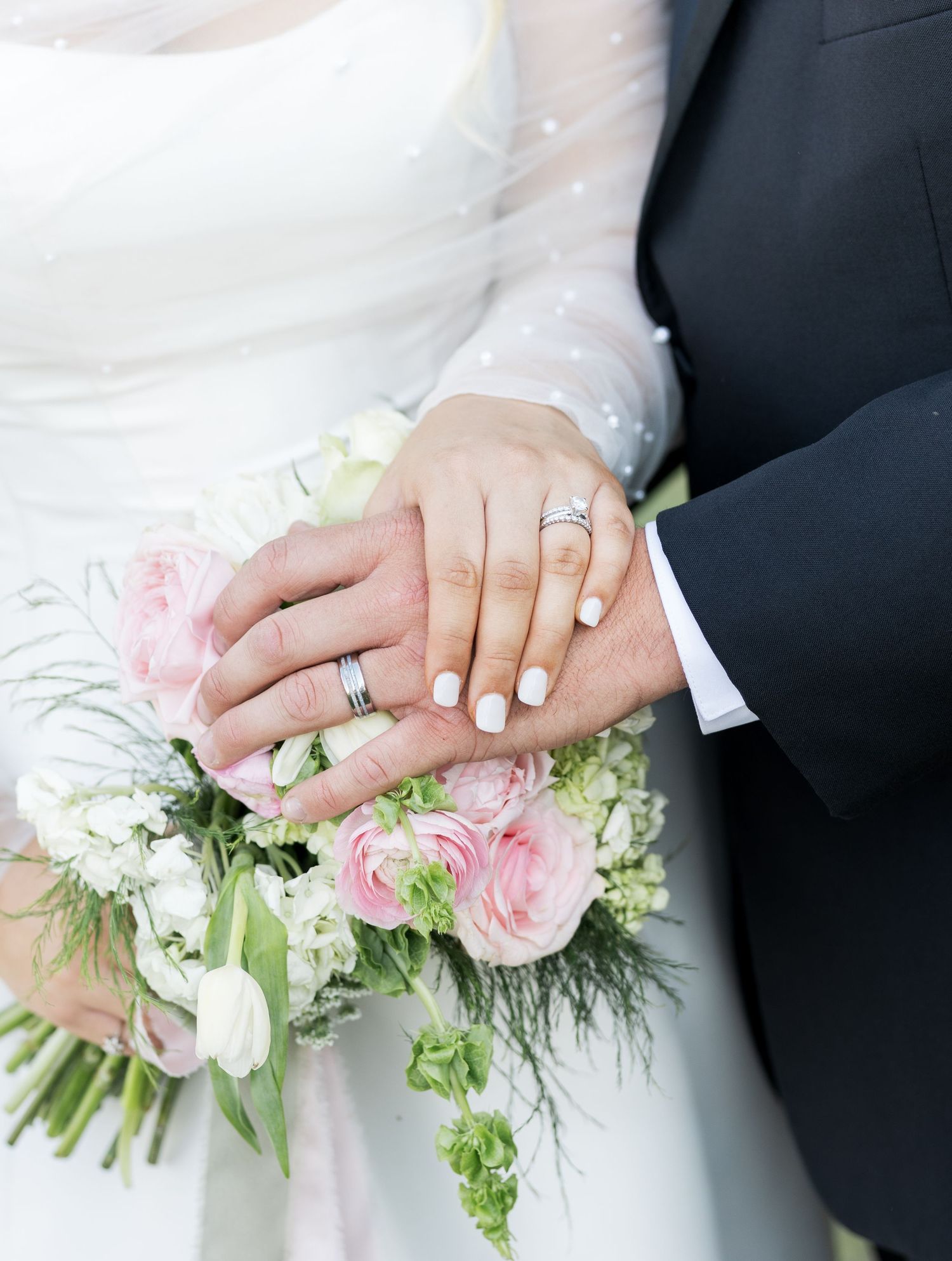 Newlyweds displaying wedding rings on hands resting over pink and white rose bridal bouquet.