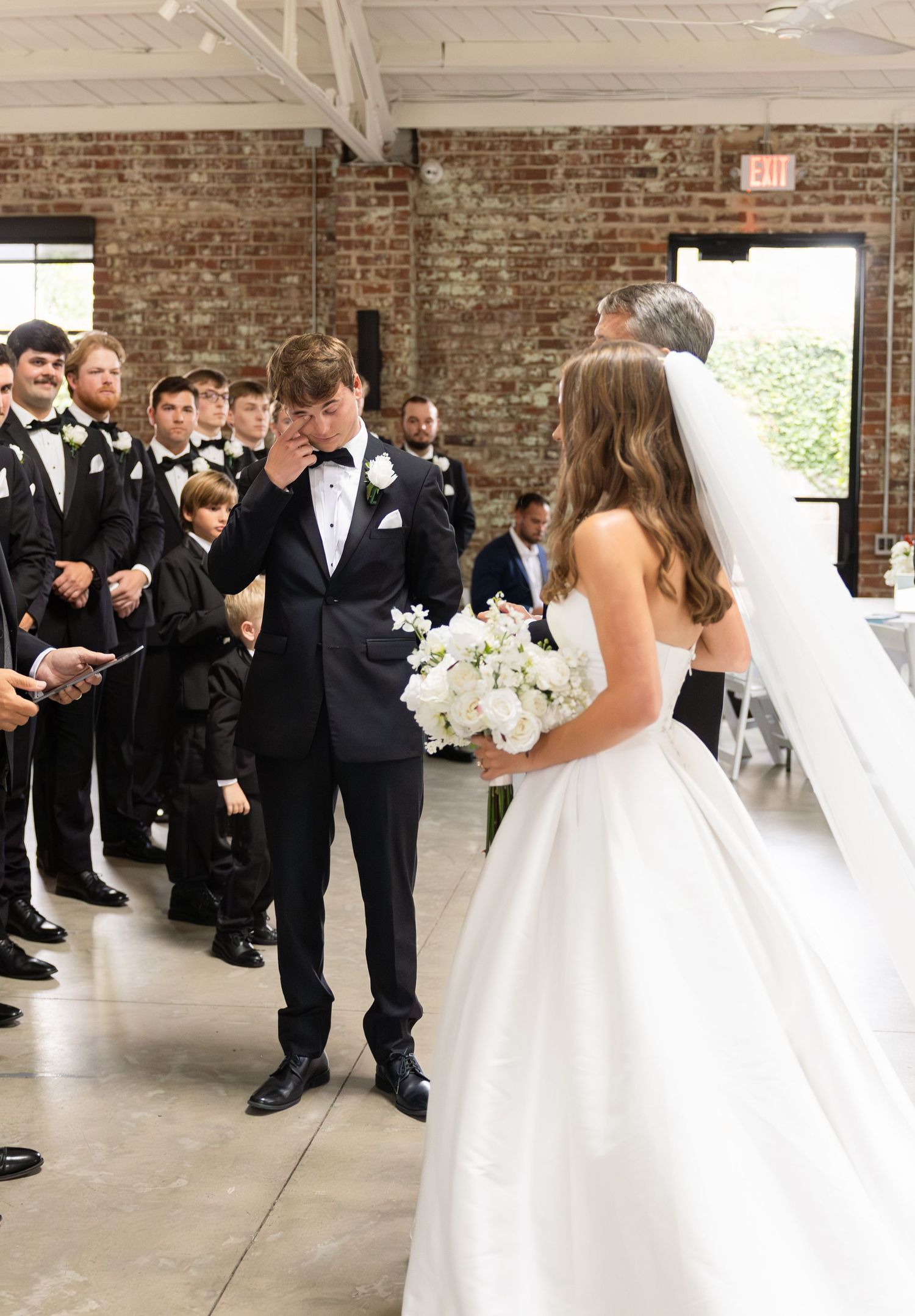 Emotional groom wiping tears as bride walks down the aisle in a white strapless gown and veil.