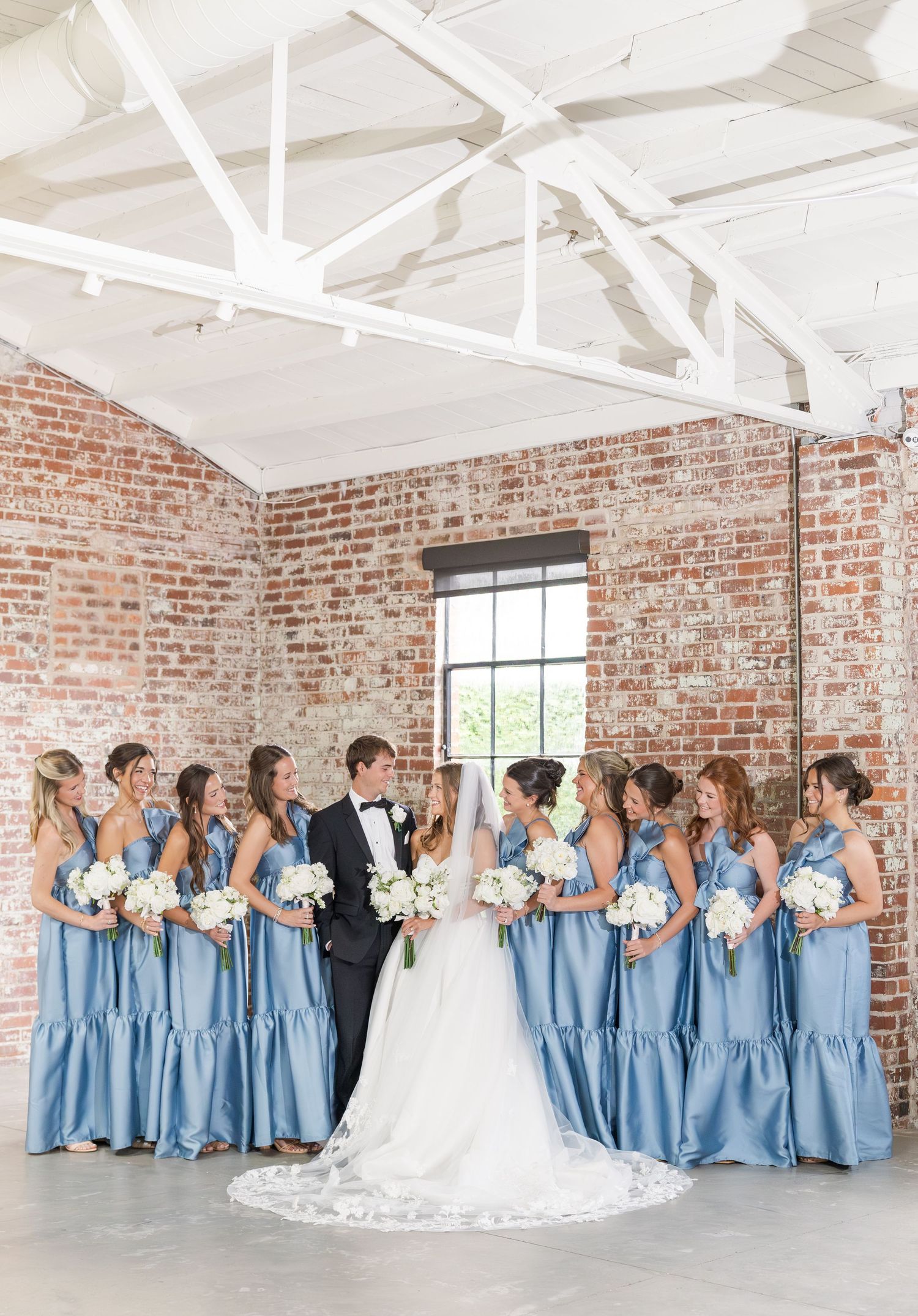 Bride and groom surrounded by bridesmaids in dusty blue gowns holding white bouquets in a rustic brick venue.