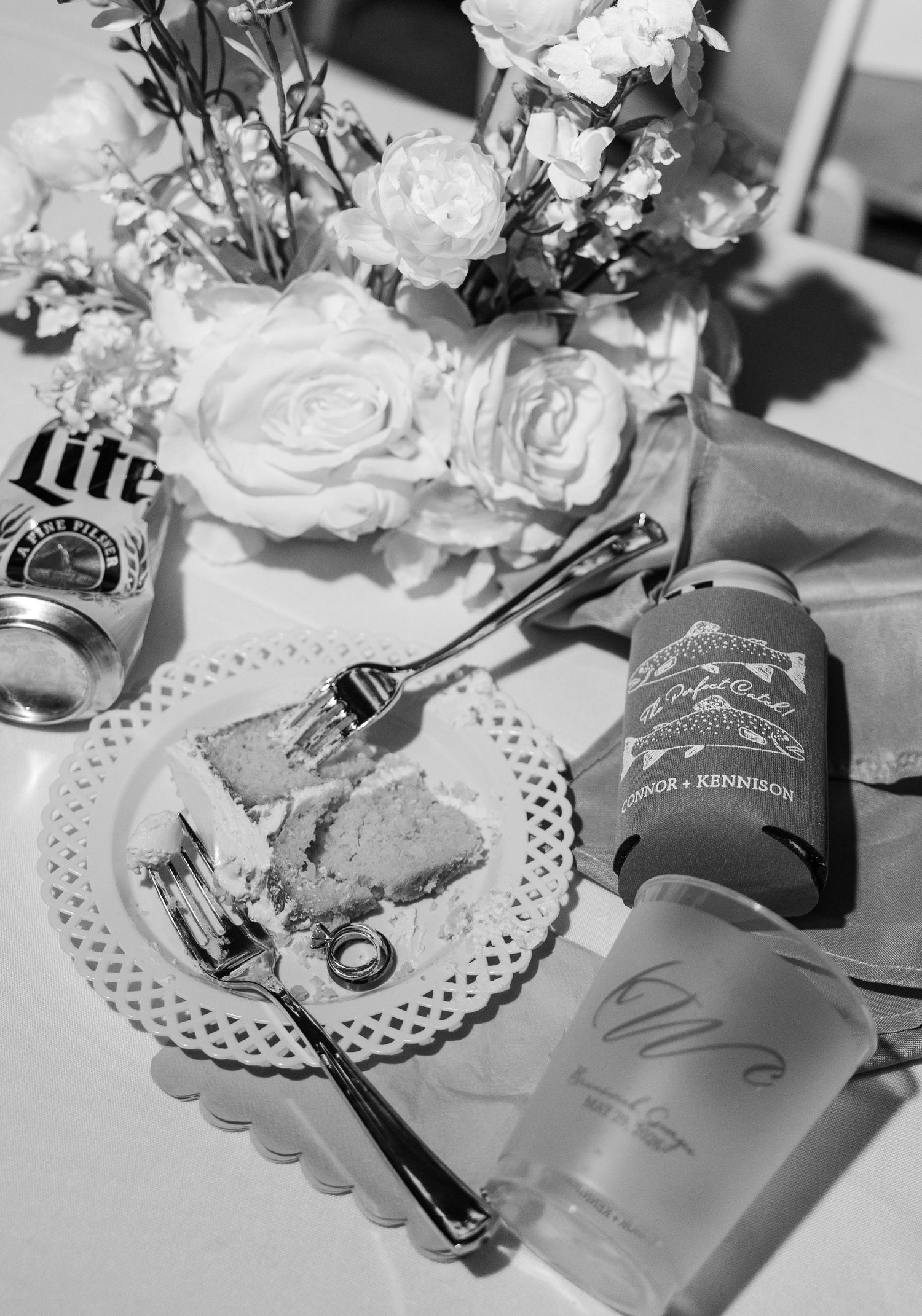 Black and white photo of wedding table with flowers, cake slice, beer can, and custom koozies.