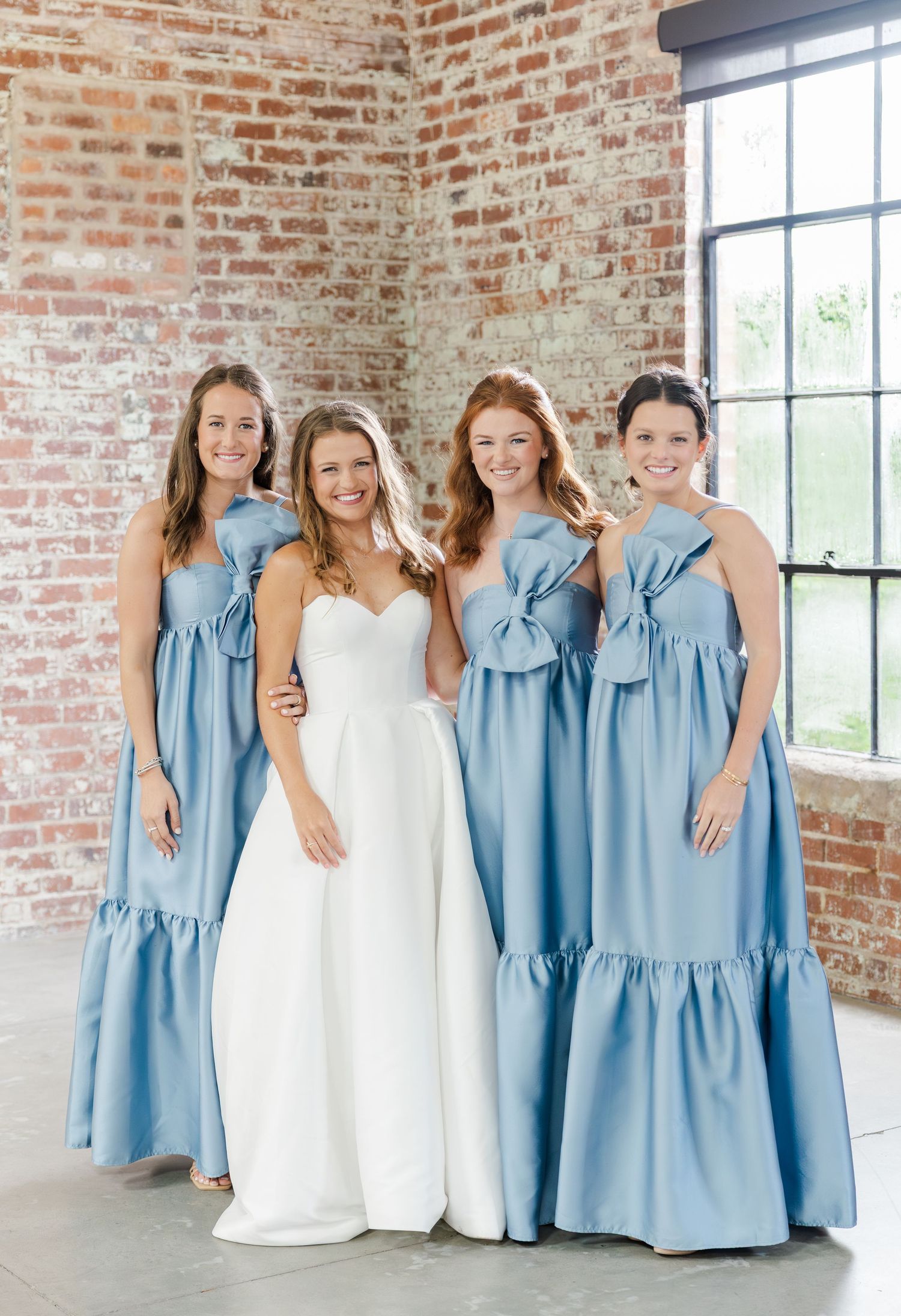 Bride in white gown poses with three bridesmaids in blue dresses against brick wall.