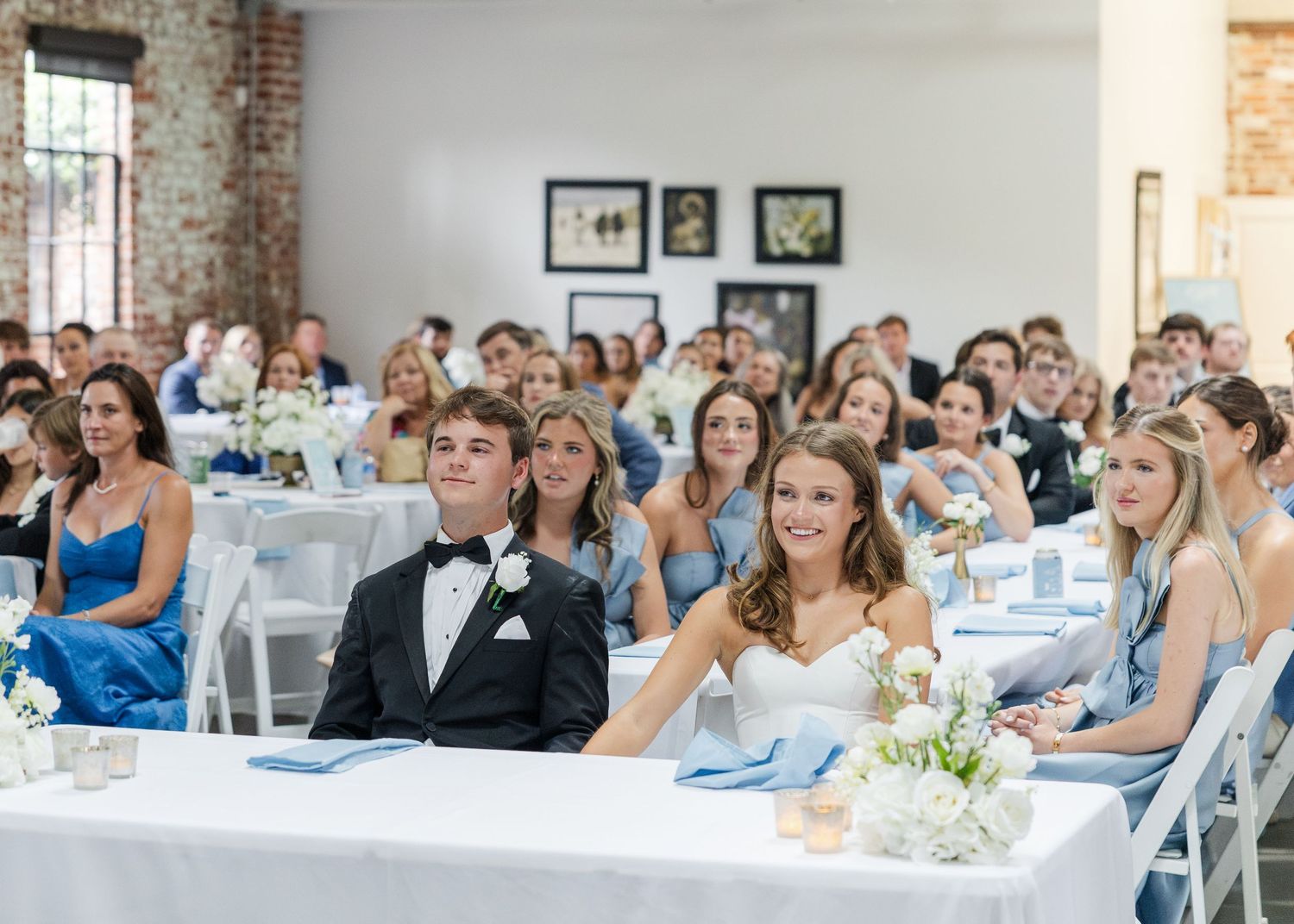 Bride and groom seated at head table smiling during wedding reception with guests in blue and white decor.