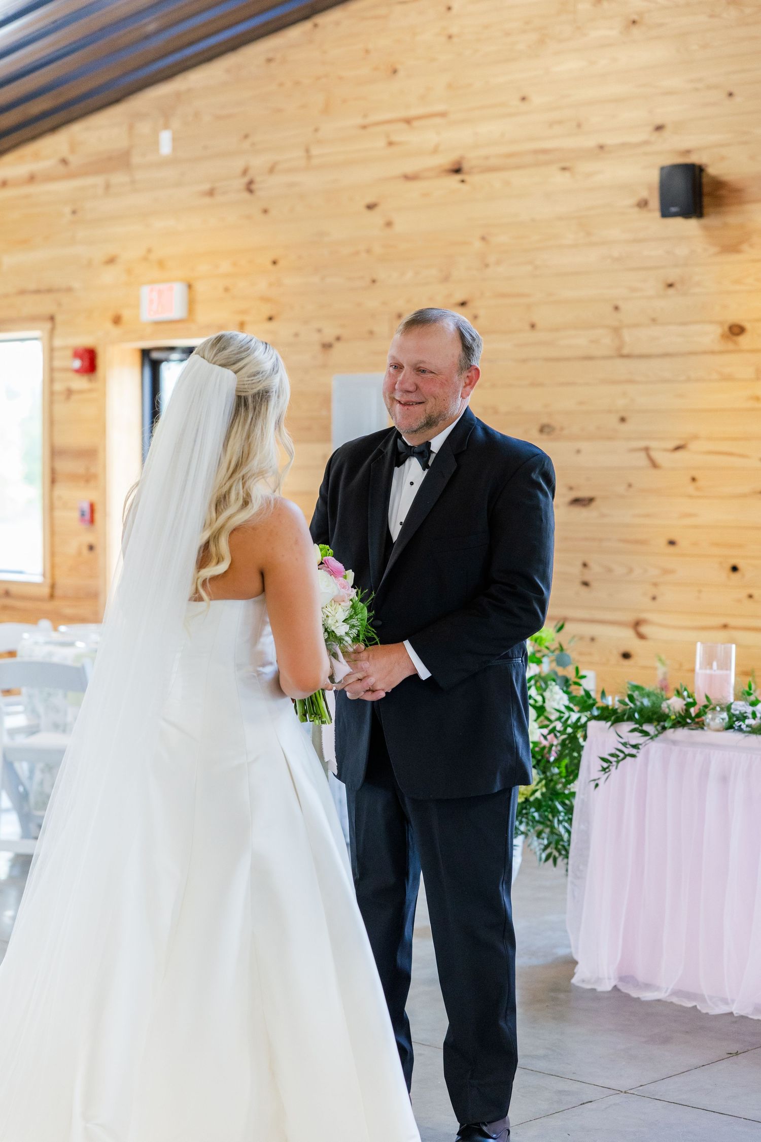 Father smiling at bride in white wedding gown during first look moment at rustic venue.