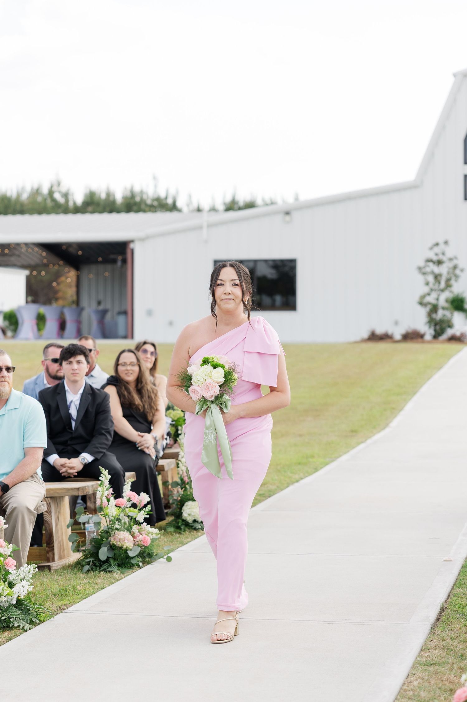 Bridesmaid in pink one-shoulder jumpsuit walks down outdoor wedding aisle carrying white and green bouquet.