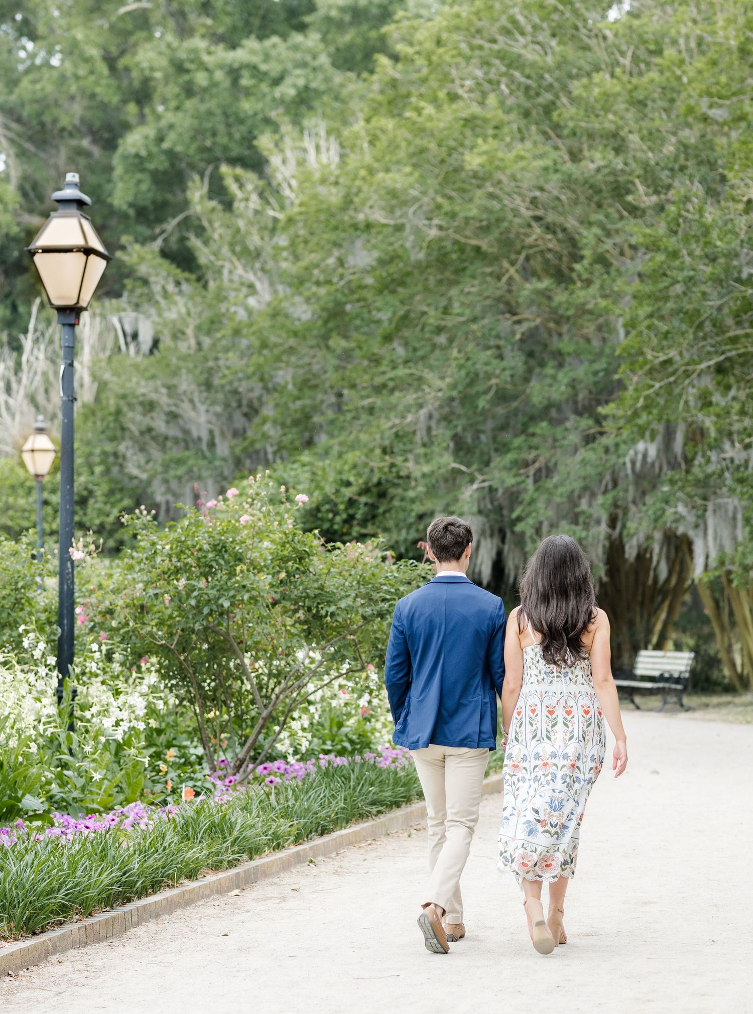 Couple walking hand-in-hand down a scenic garden path lined with lamp posts and lush greenery.