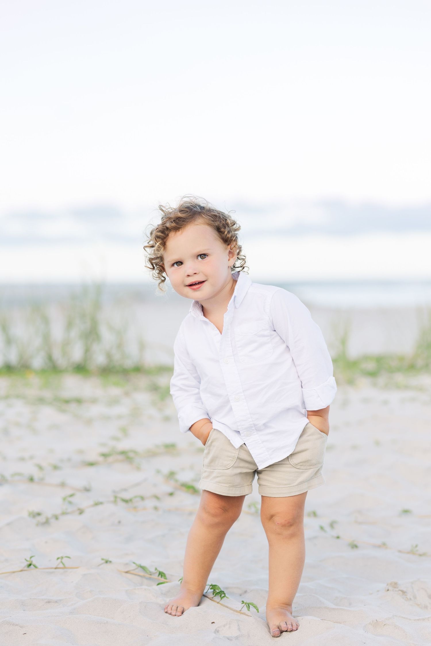 A young child in a white shirt and khaki shorts stands playfully on a beach.