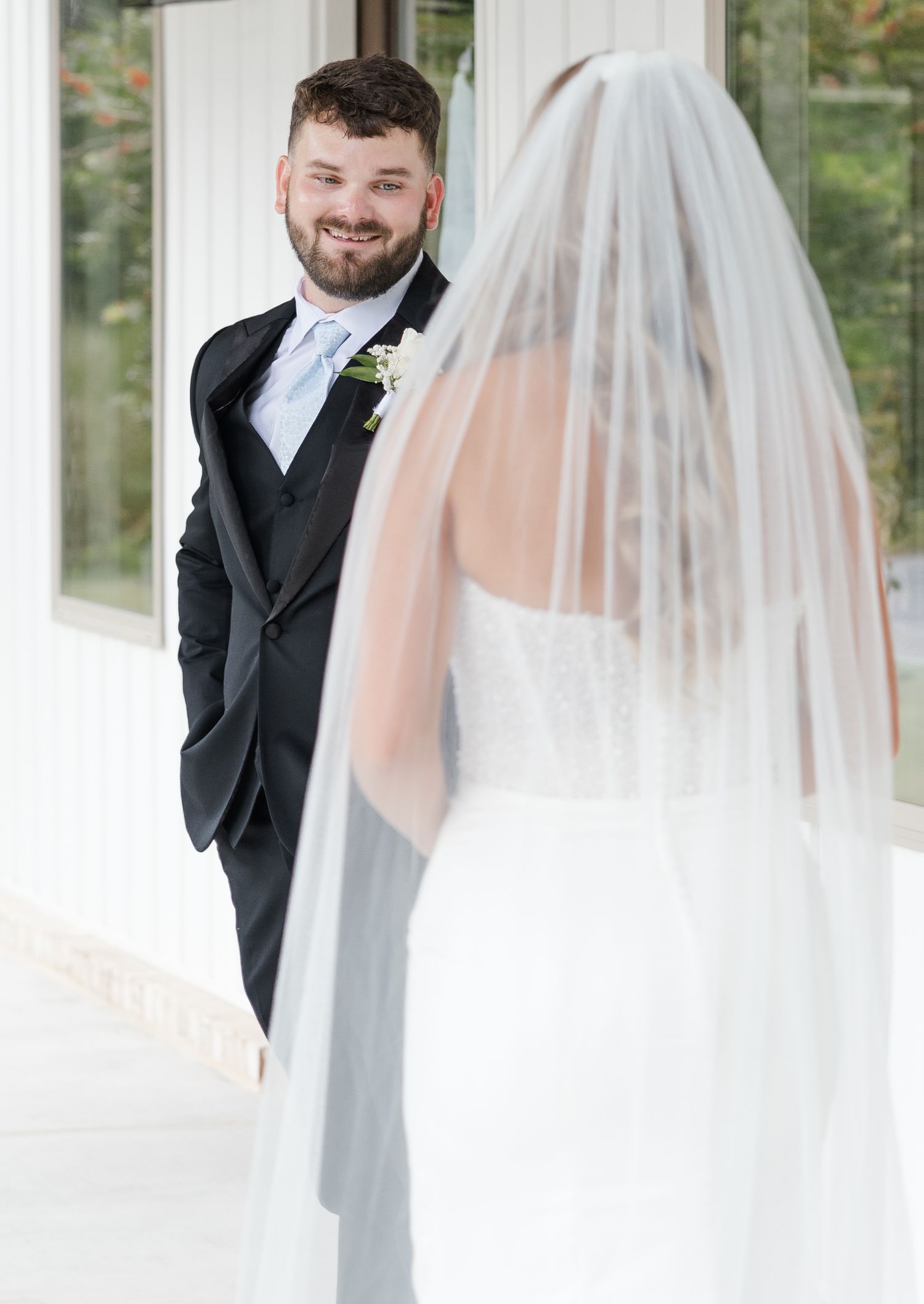 Groom in black tuxedo smiles joyfully at his veiled bride during an emotional first look moment at their wedding.