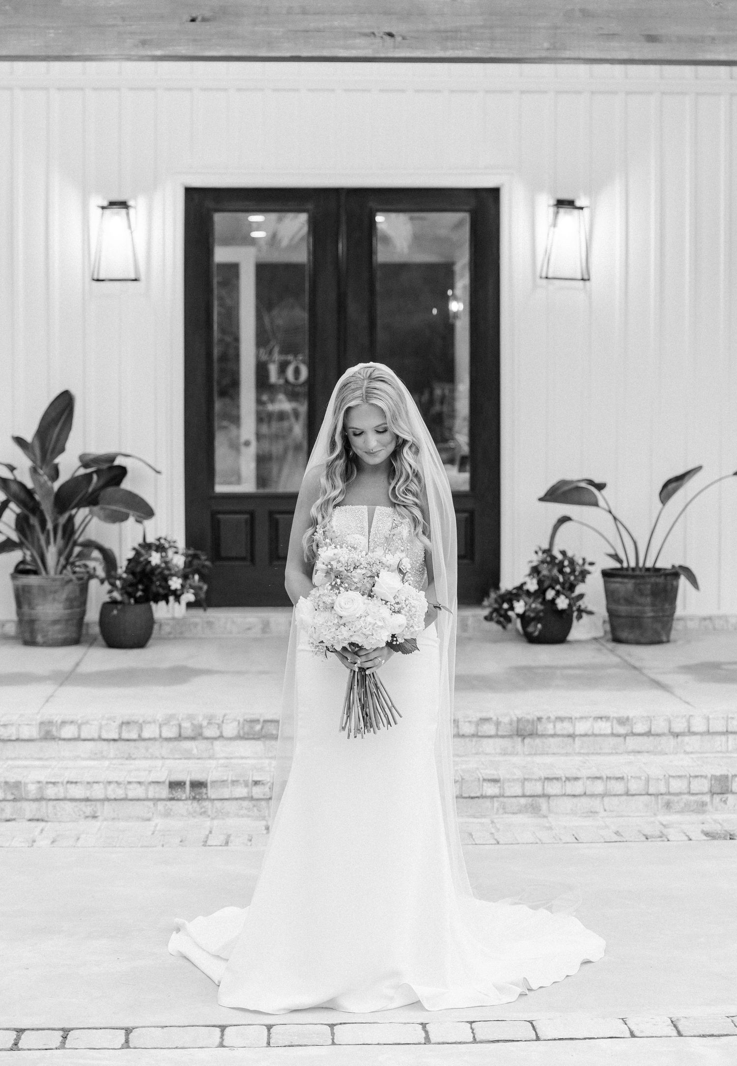 Black and white photo of a bride in a flowing gown and veil holding a bouquet outside elegant venue doors.