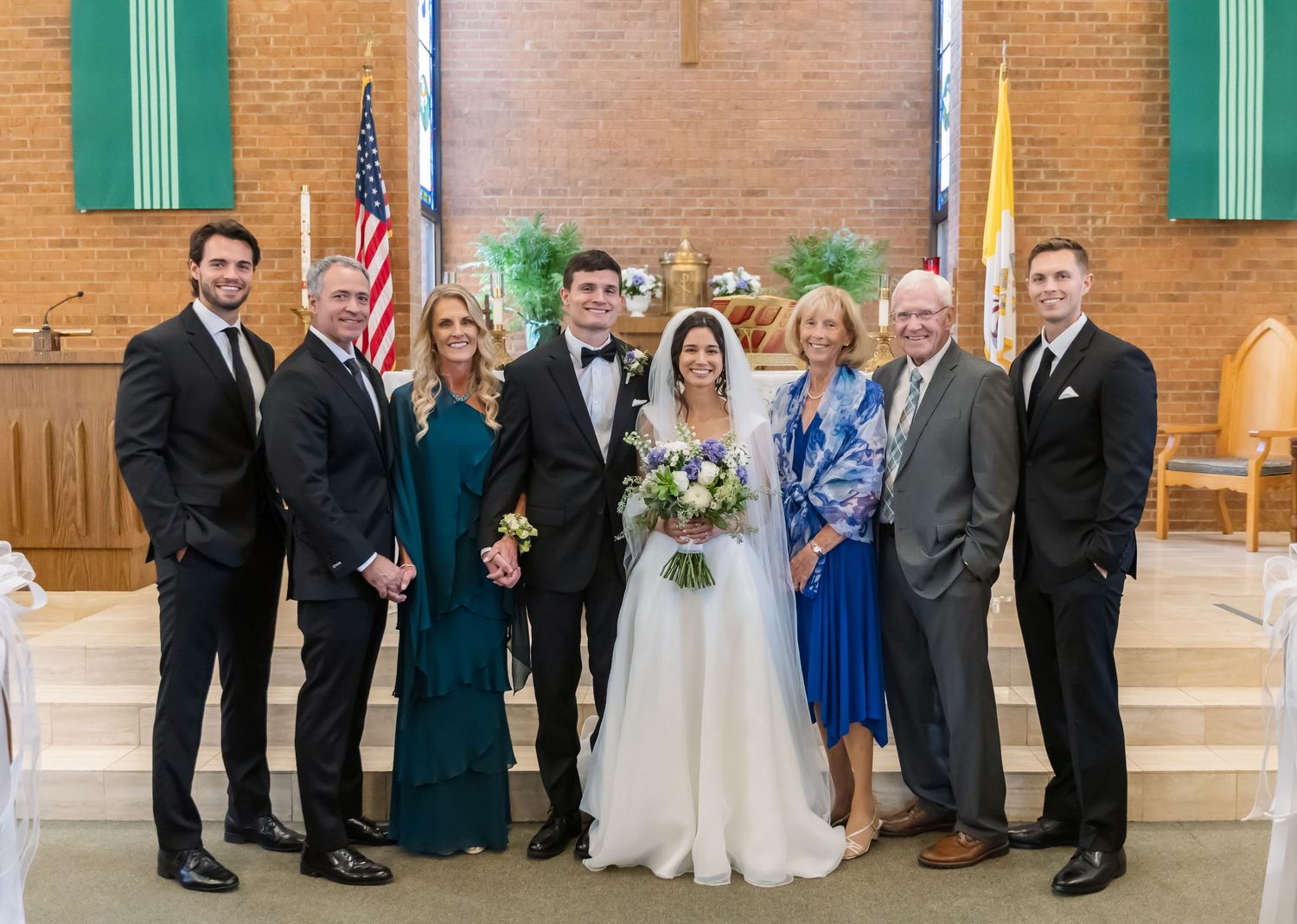 A panoramic wedding party photo in a church with exposed brick walls and teal accents.