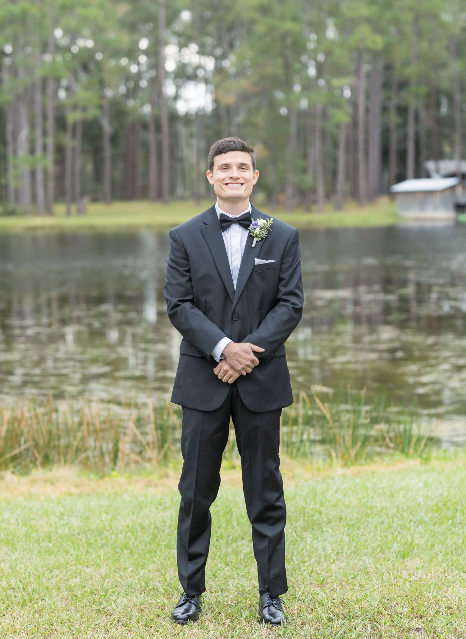 A formal outdoor portrait series showing someone in a black tuxedo standing by a lake with trees in the background.