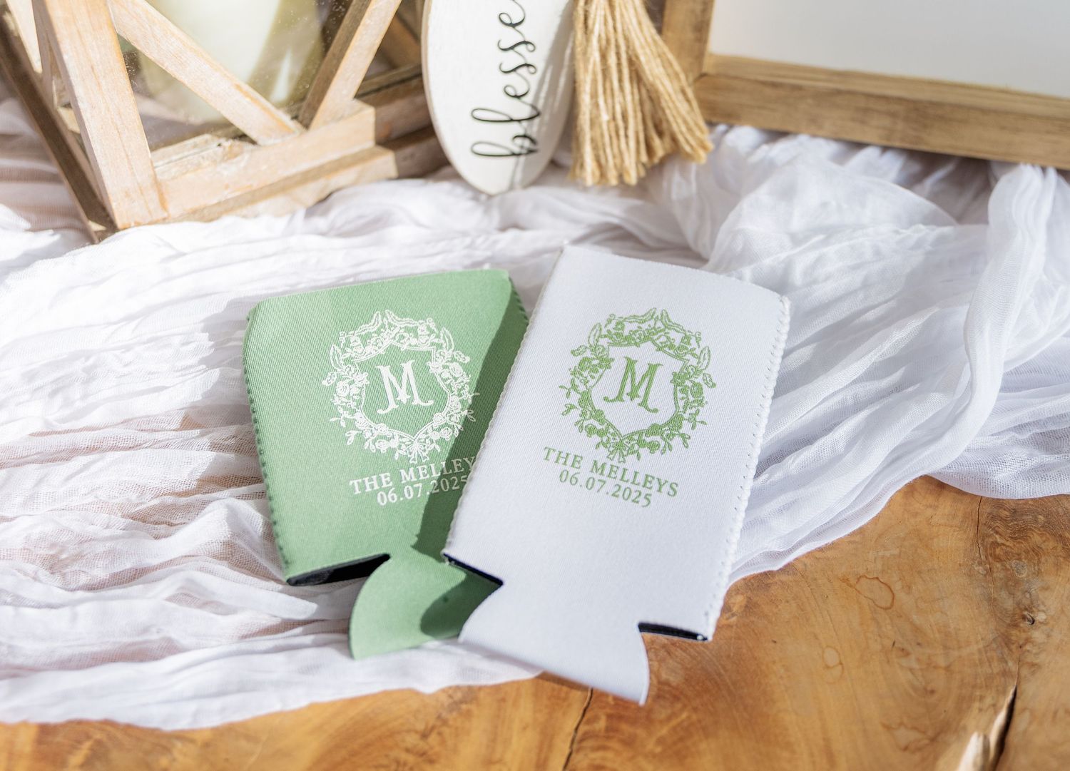 Mint green and white monogrammed wedding koozies with floral wreath design displayed on white fabric.