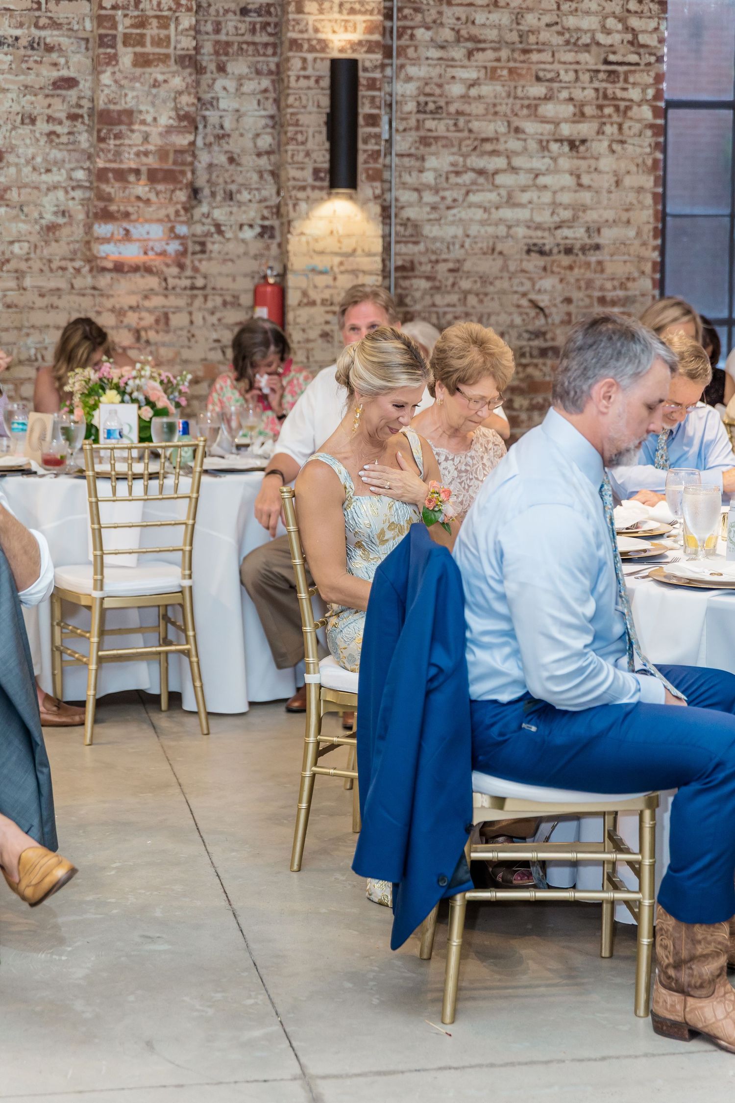 Wedding reception guests seated at round tables with white linens in a rustic brick venue.