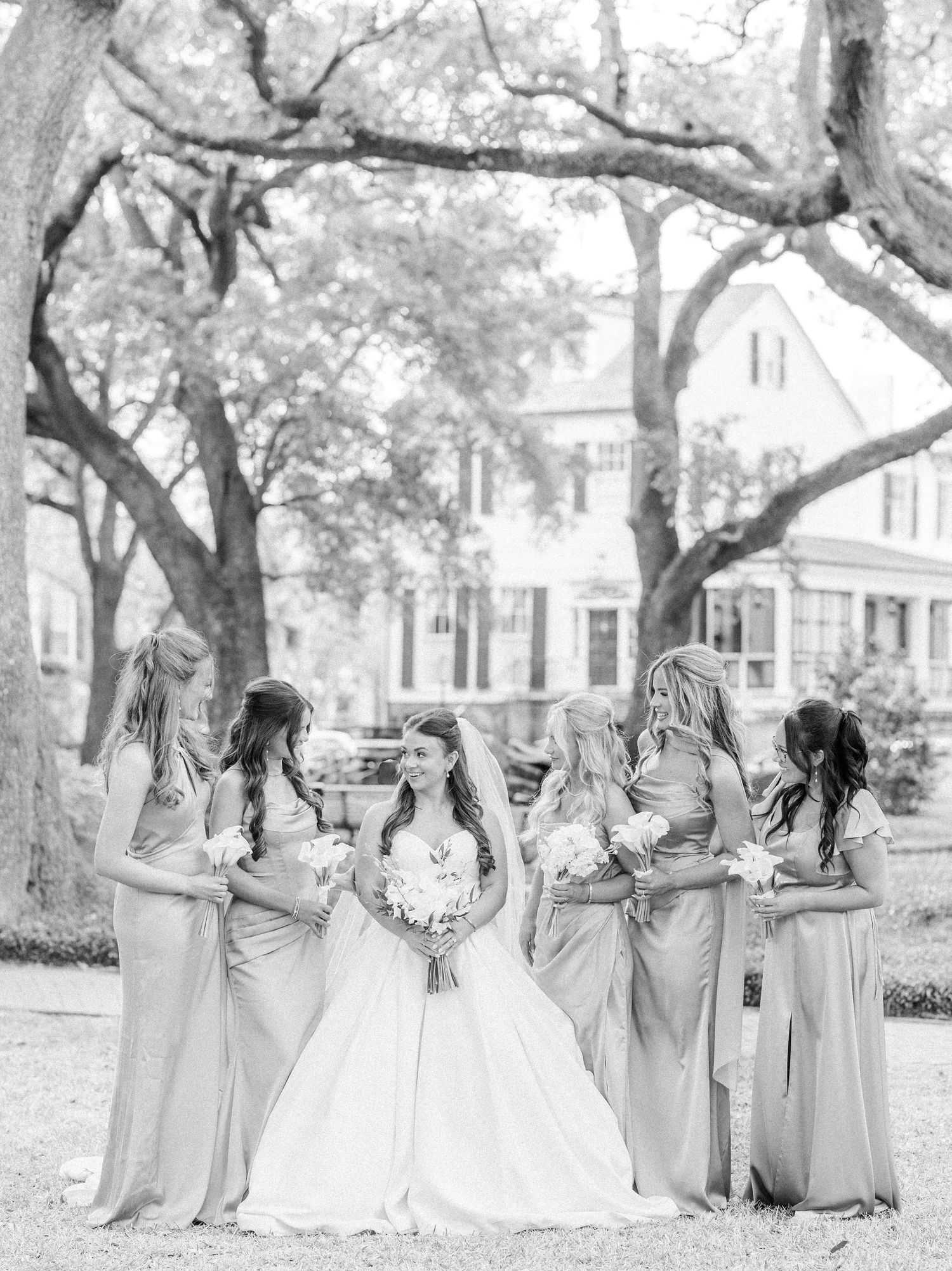Wedding party in light gray dresses poses together under oak trees at an outdoor venue.