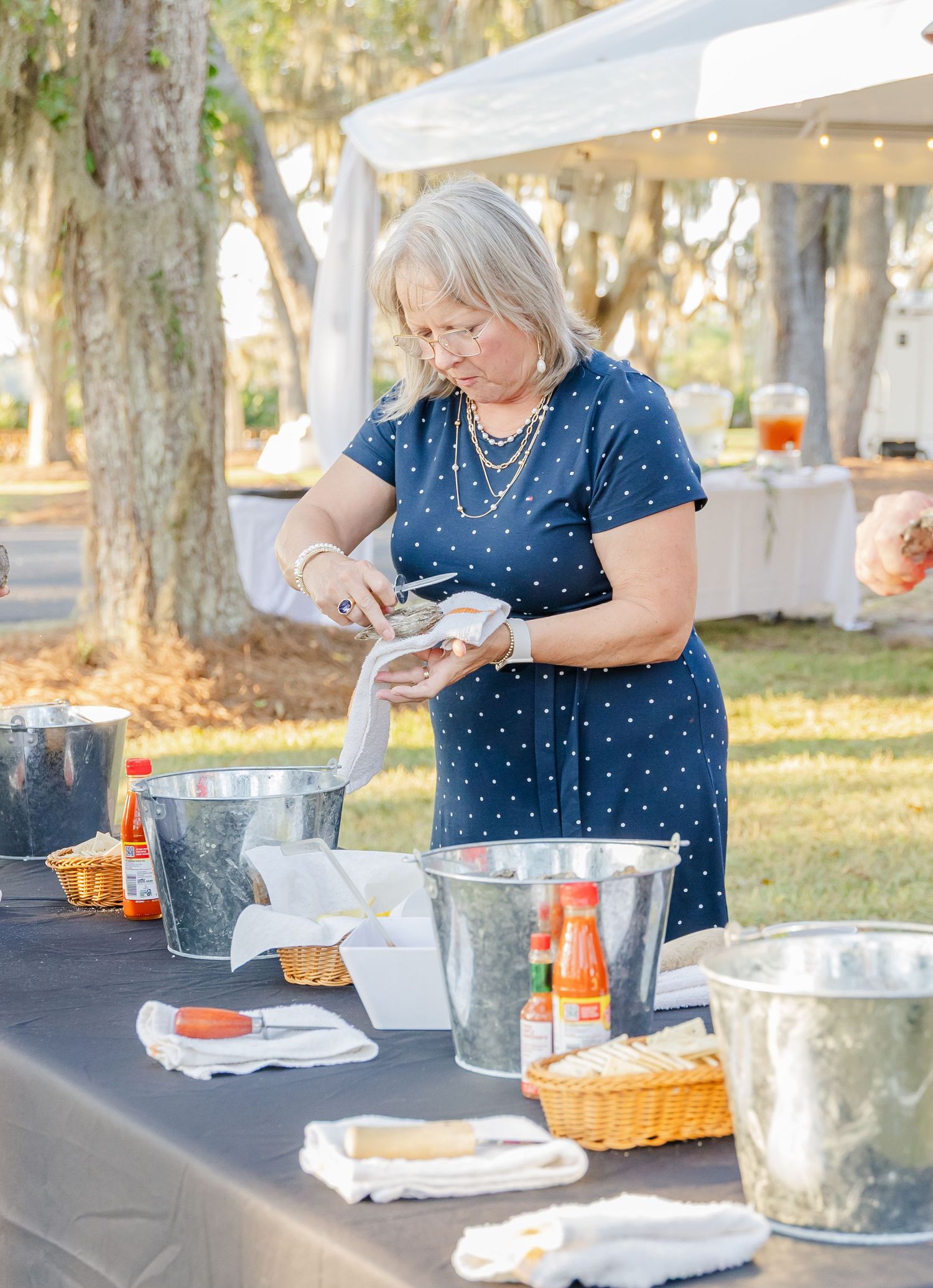 A person in a polka dot blue dress serves food at an outdoor buffet table under a white tent.
