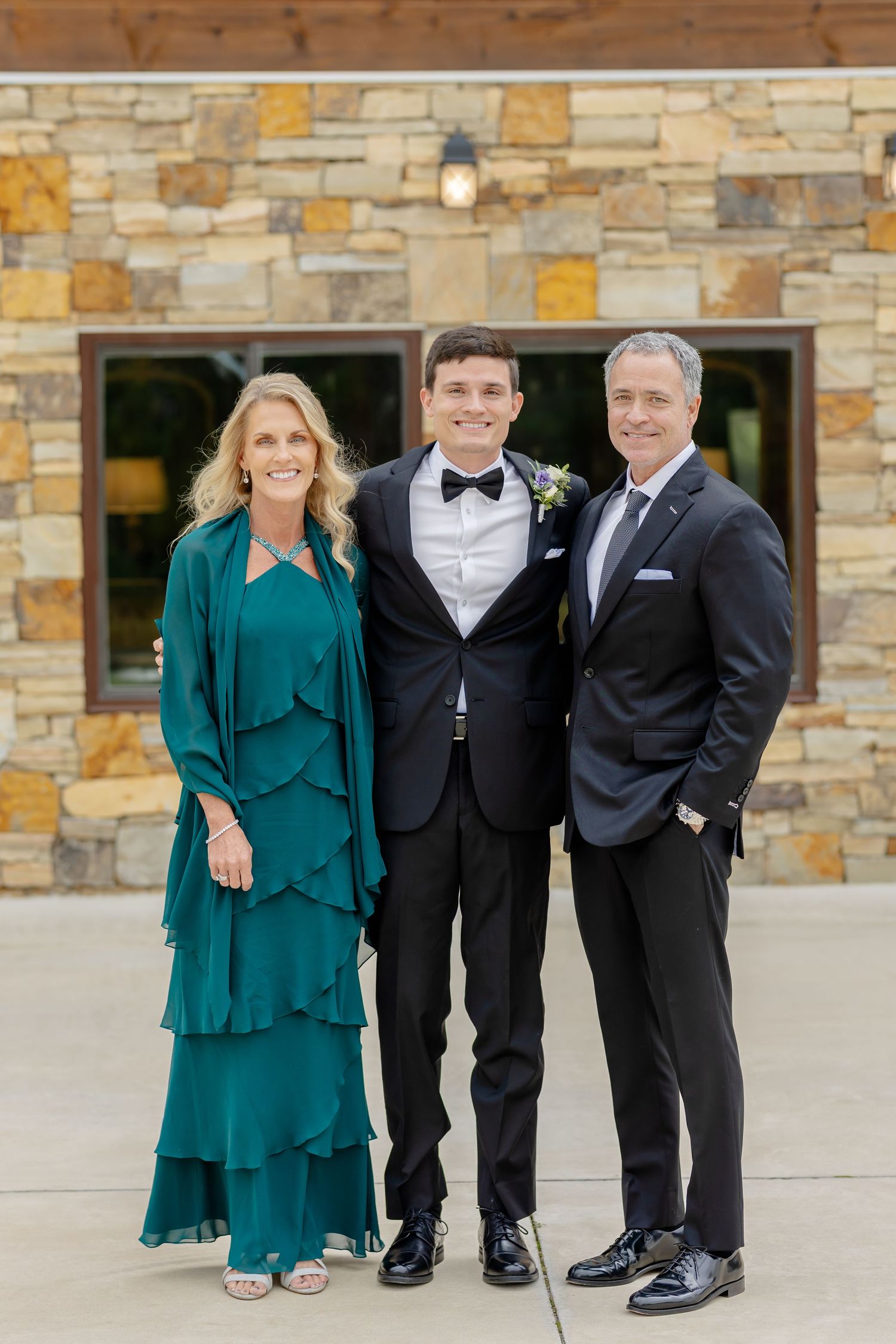 A group of people wearing formal attire pose together against a stone wall backdrop.