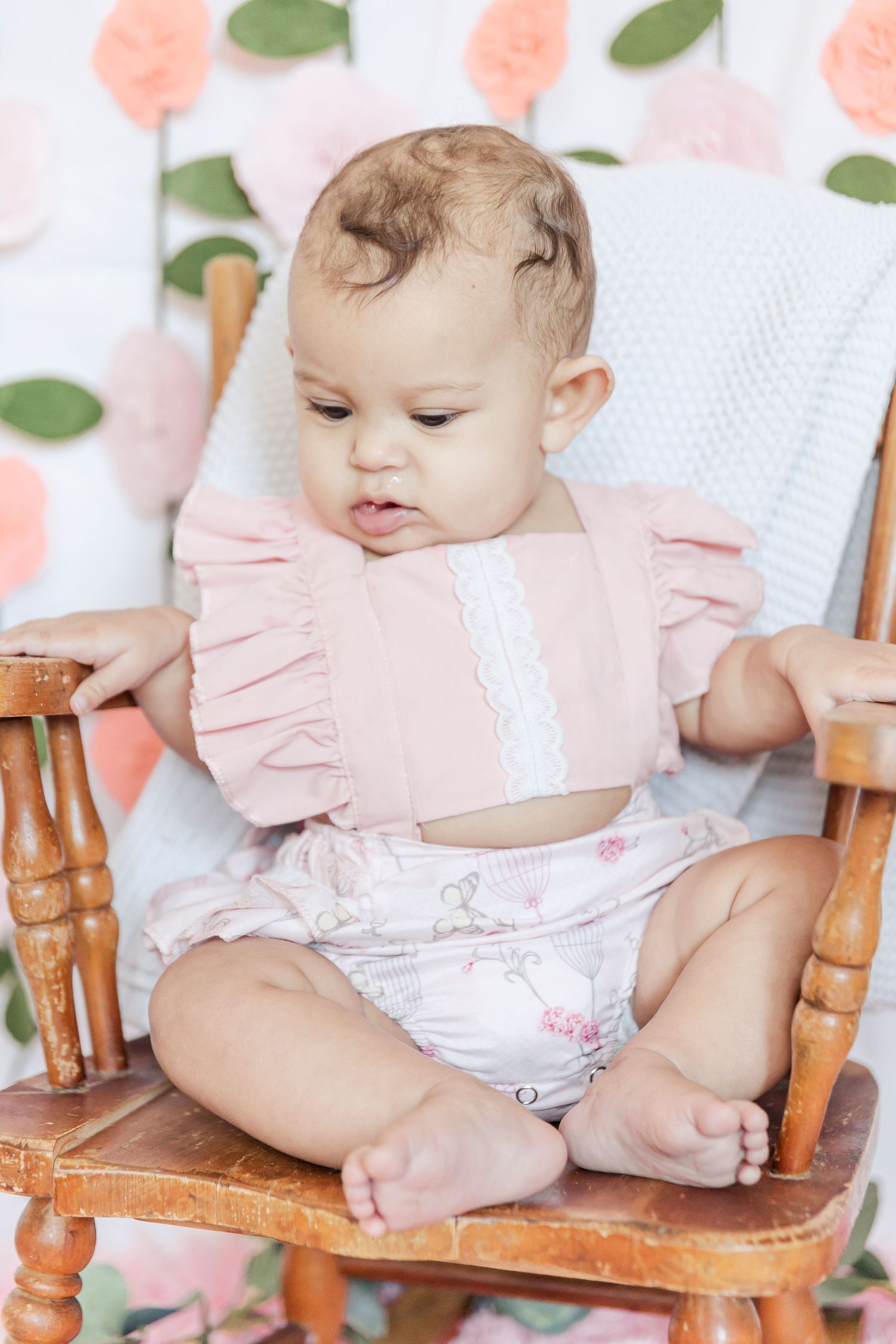 Baby sitting in wooden chair wearing pink ruffled outfit against floral backdrop.