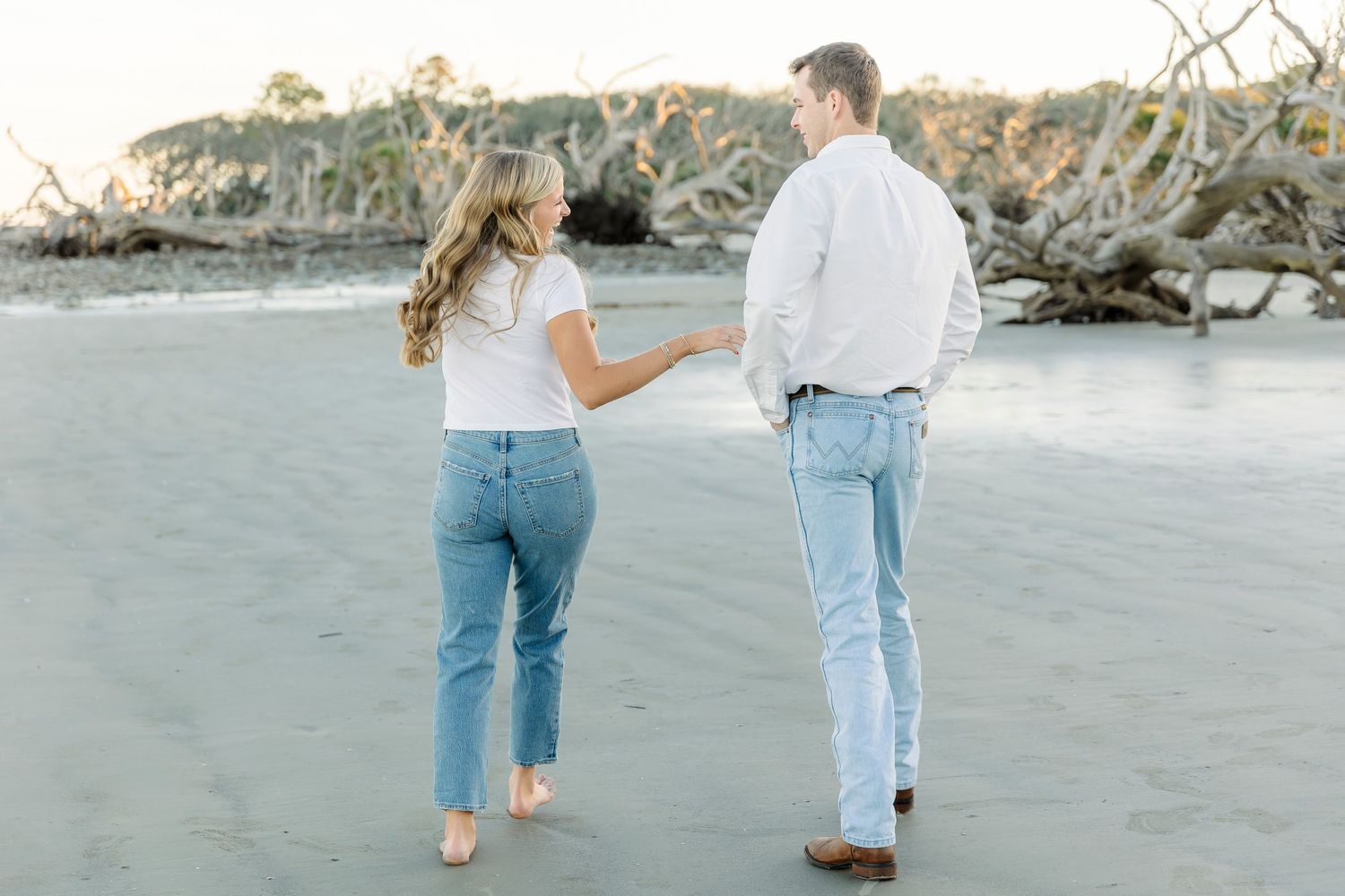 Couple in matching white tops and jeans walking barefoot on a beach with driftwood in background.