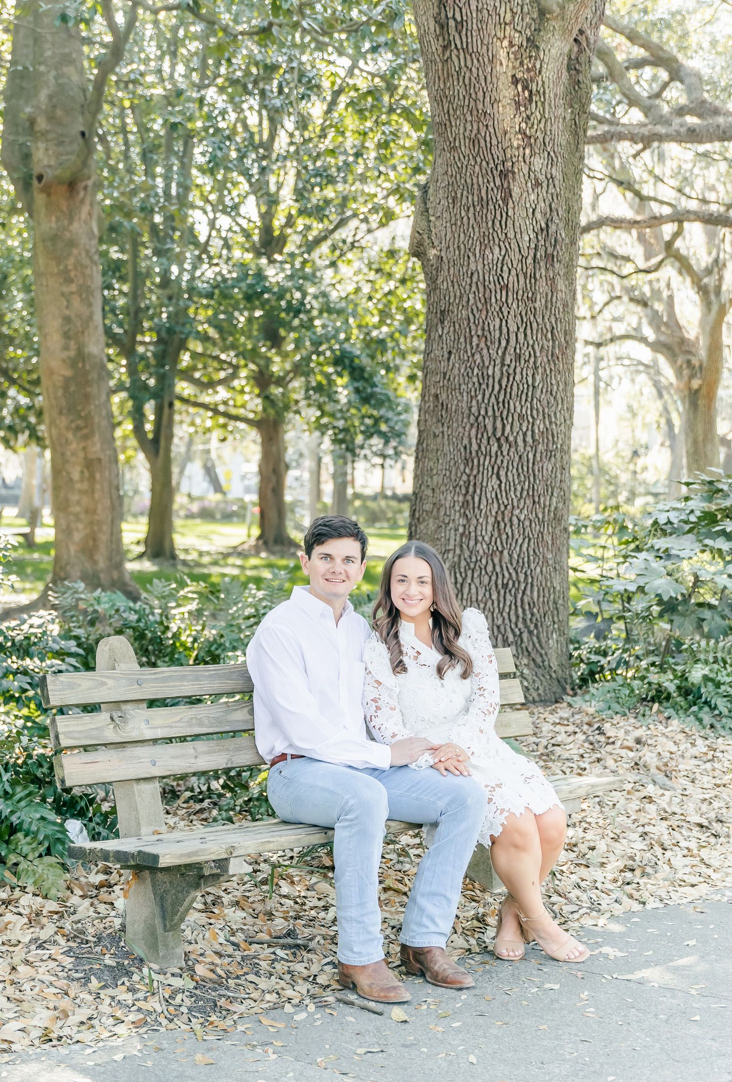Engaged couple sitting together on wooden bench beneath large oak tree in garden setting.
