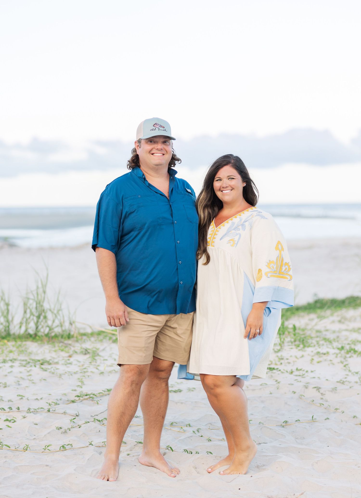 A couple poses on the beach wearing casual attire against an ocean background.