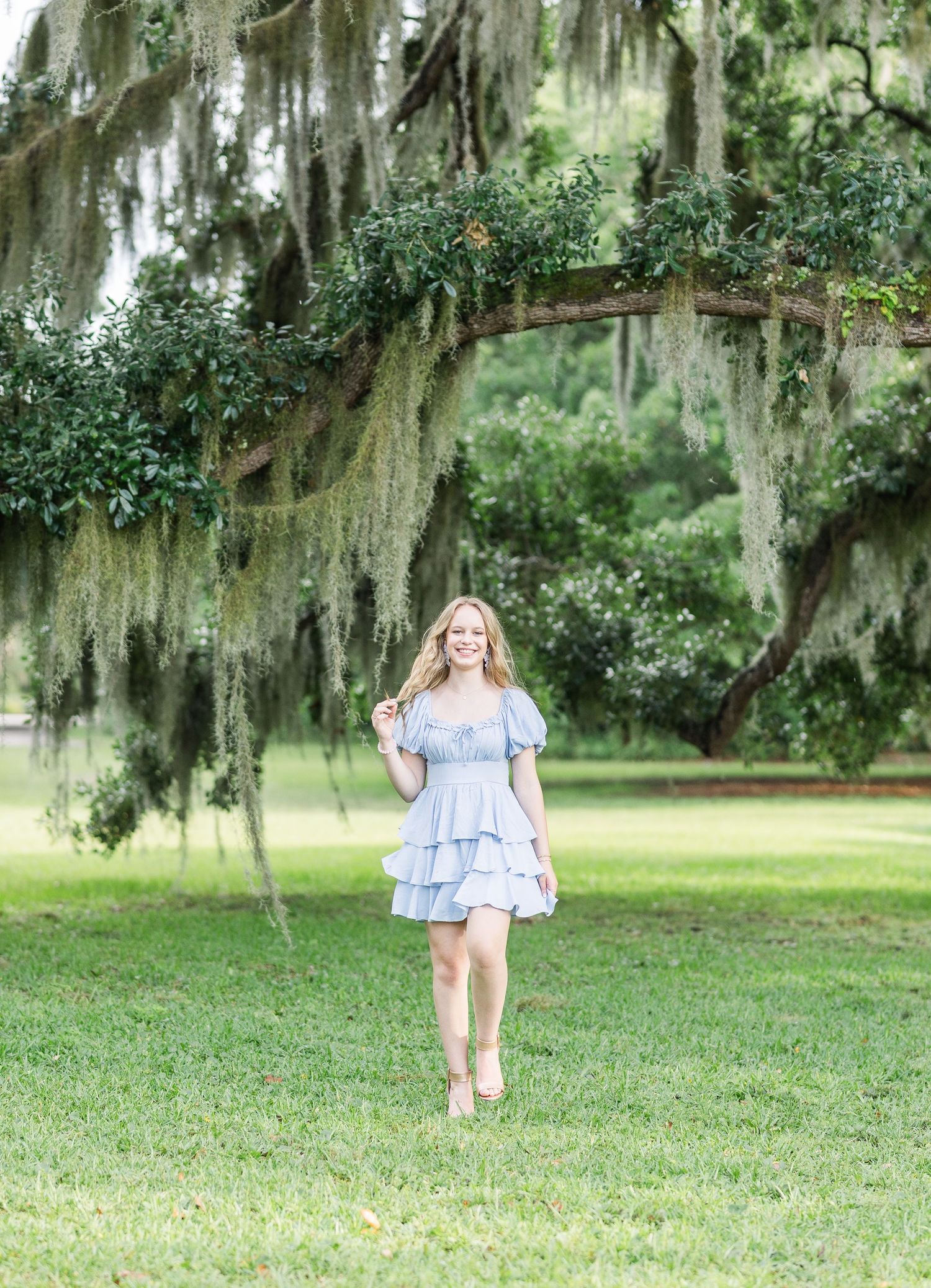 Person walking through scenic southern garden with Spanish moss hanging from oak trees on sunny day.