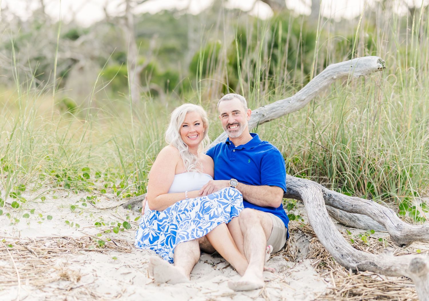 Couple in coordinated blue and white outfits sitting by driftwood on beach with tall grass in background.