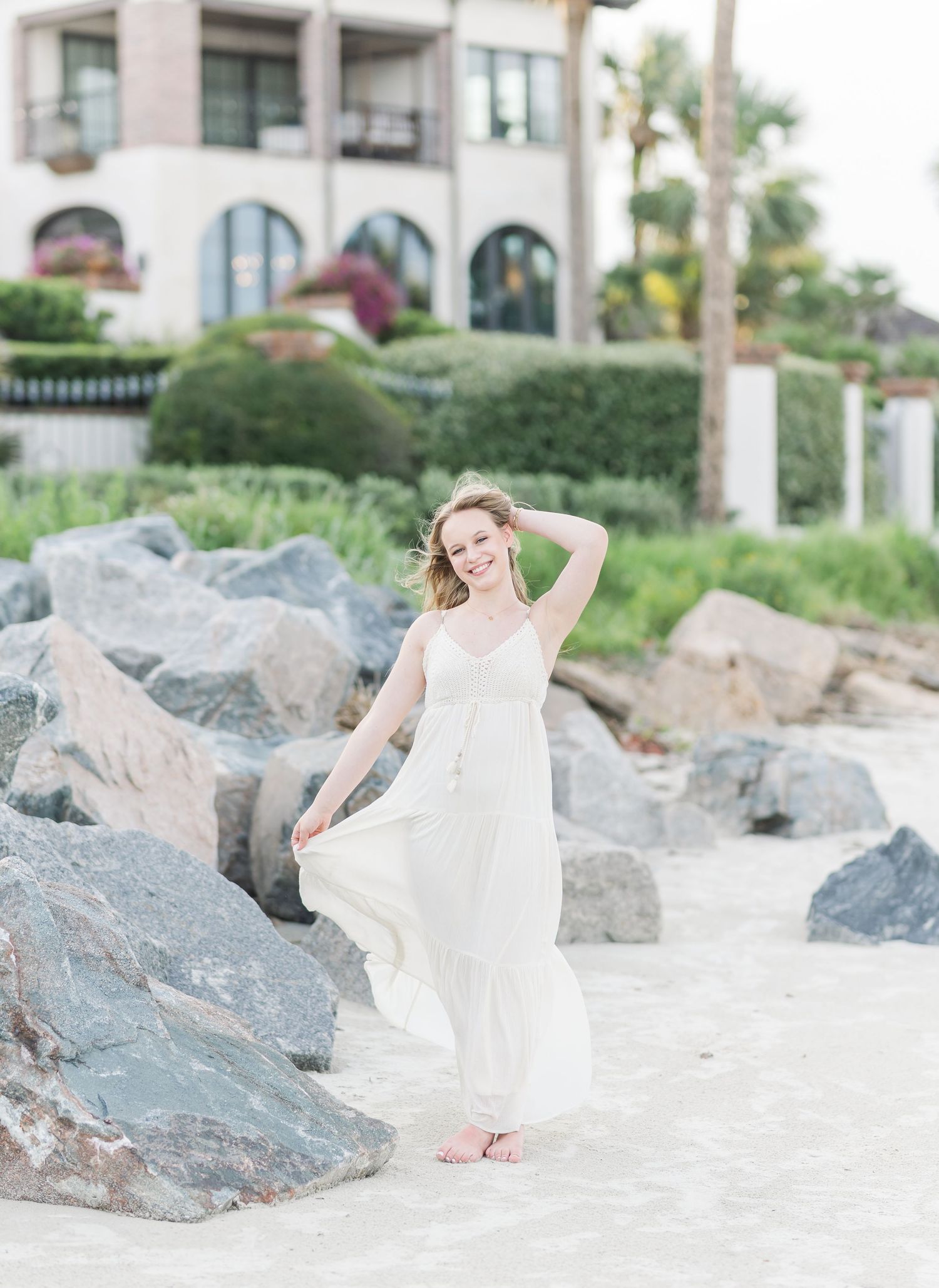 Beachside portrait series near large rocks featuring elegant poses in a white flowing dress.