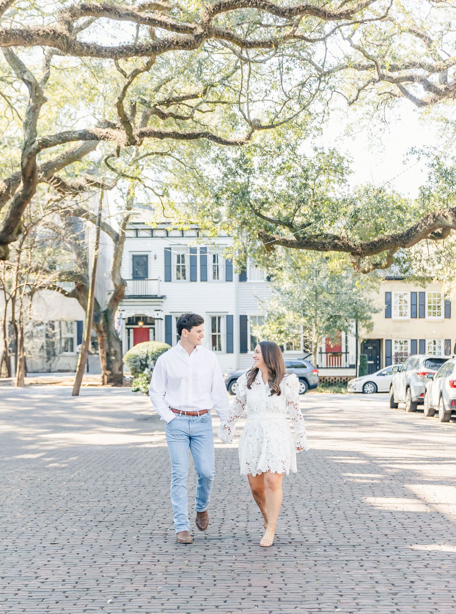 A couple walks hand in hand along a brick street lined with charming historic homes and oak trees.