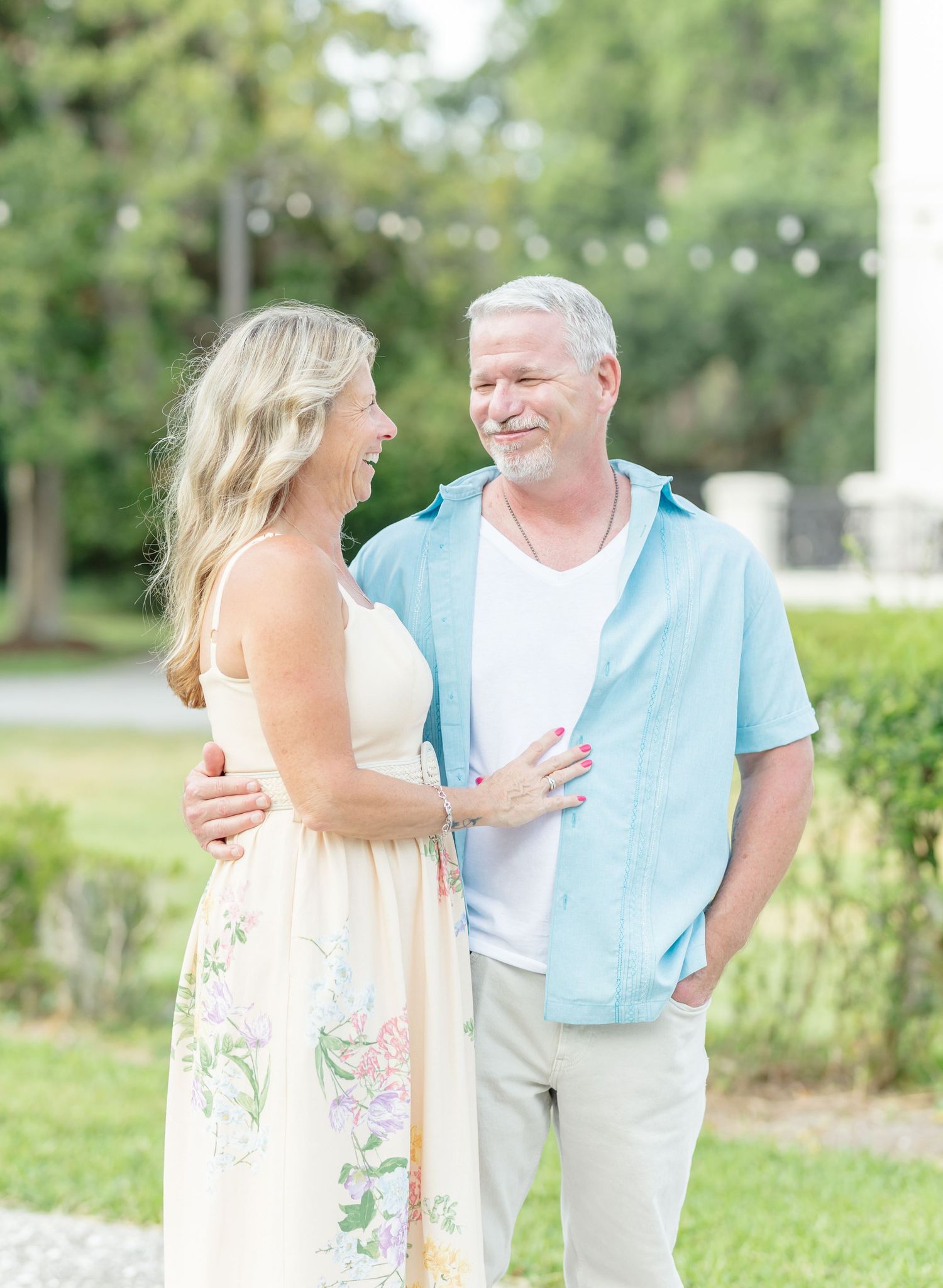 A couple in casual summer attire stands together in a lush garden setting with green foliage in the background.