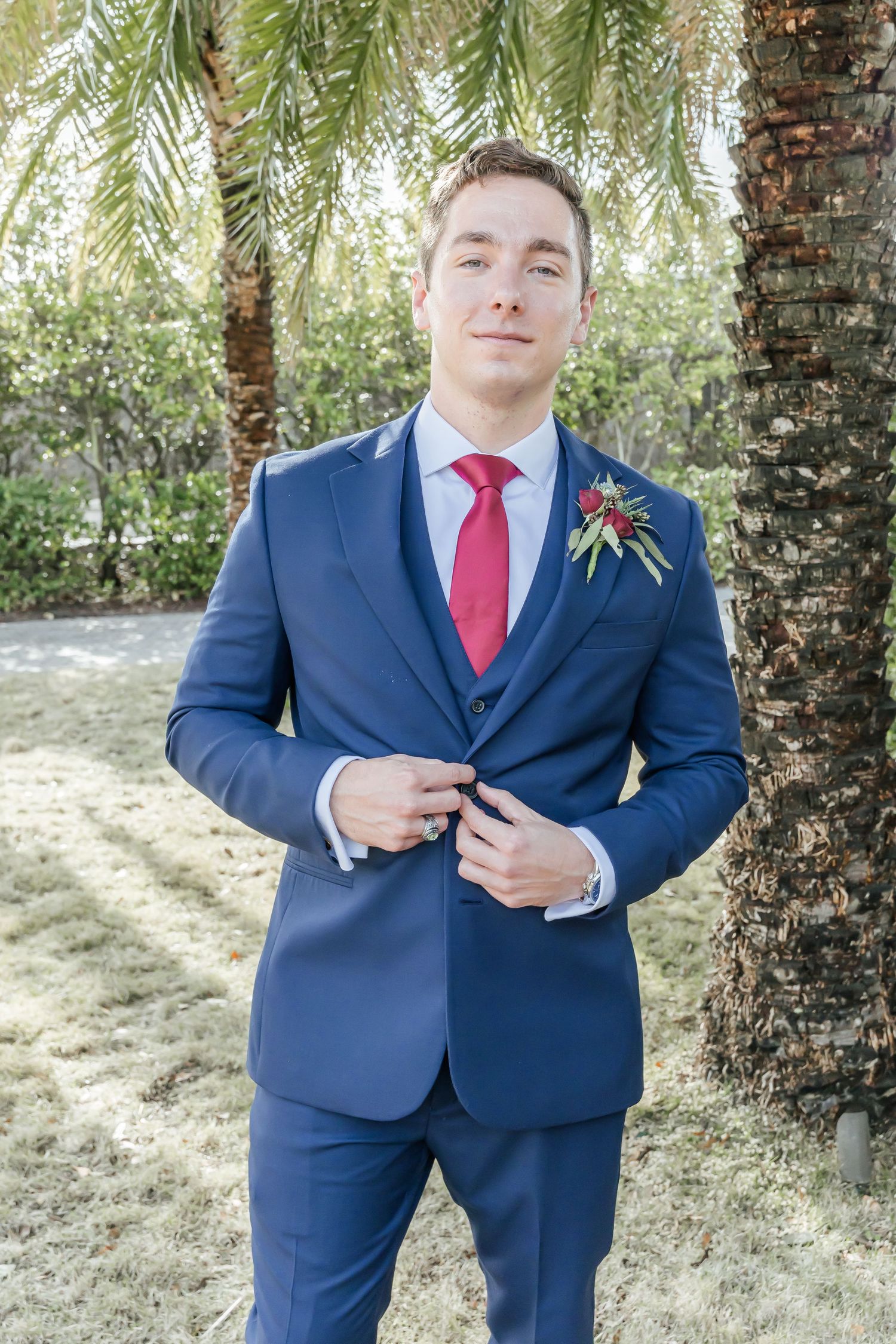Groom in navy suit and red tie adjusts his cuff links during outdoor wedding photoshoot with palm trees.
