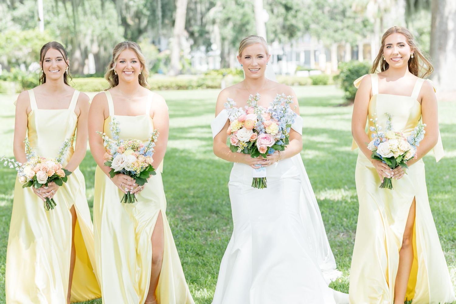 Bride in white gown with bridesmaids in yellow dresses holding spring bouquets outdoors.