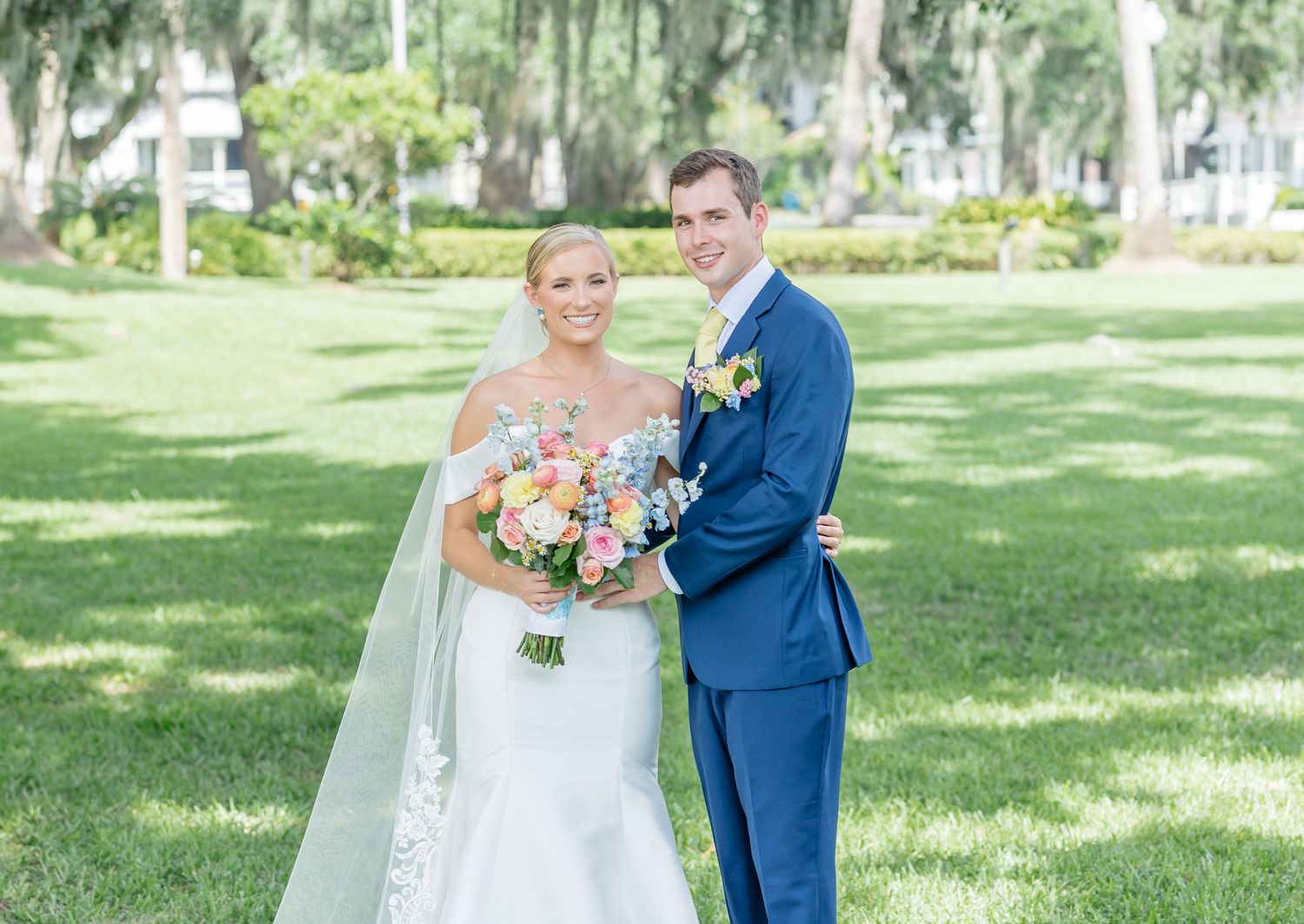 A wedding couple poses outdoors under moss-draped trees with colorful summer bouquet.