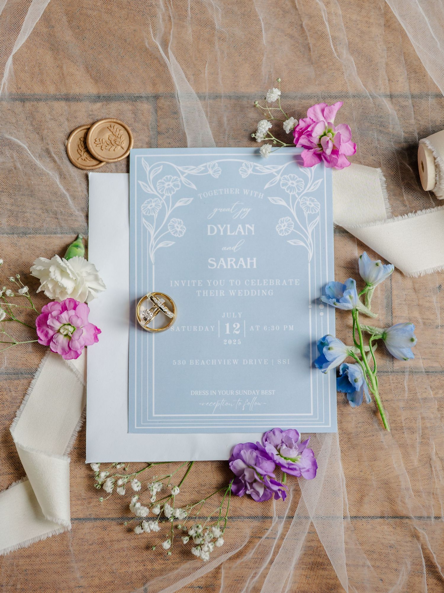 Light blue wedding invitation with floral design surrounded by pink, purple and blue flowers on brown paper background.