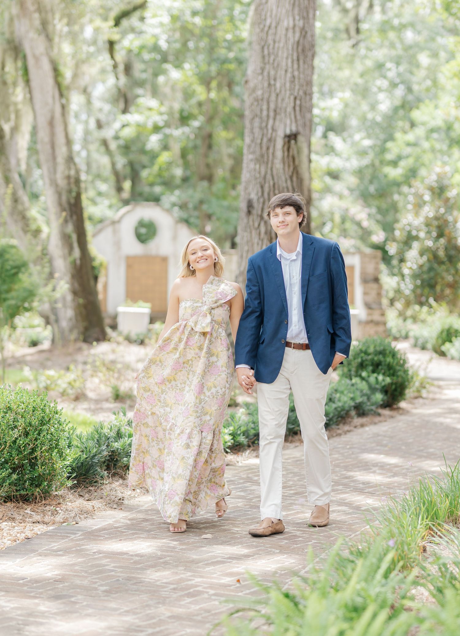 Engaged couple walking along a tree-lined garden pathway in elegant spring attire.