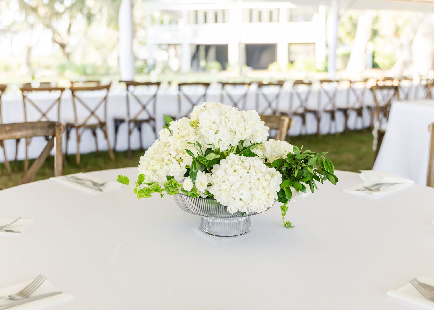 Elegant white wedding reception tables decorated with fresh floral centerpieces and natural greenery under a white tent.