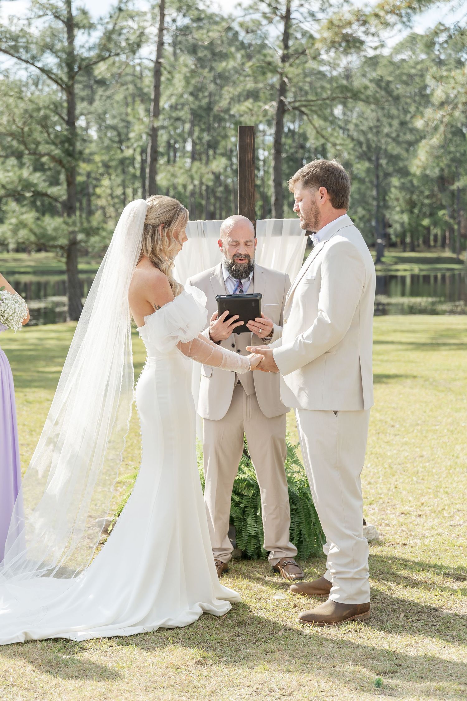 Outdoor wedding ceremony under tall pine trees with couple and officiant in light-colored attire.