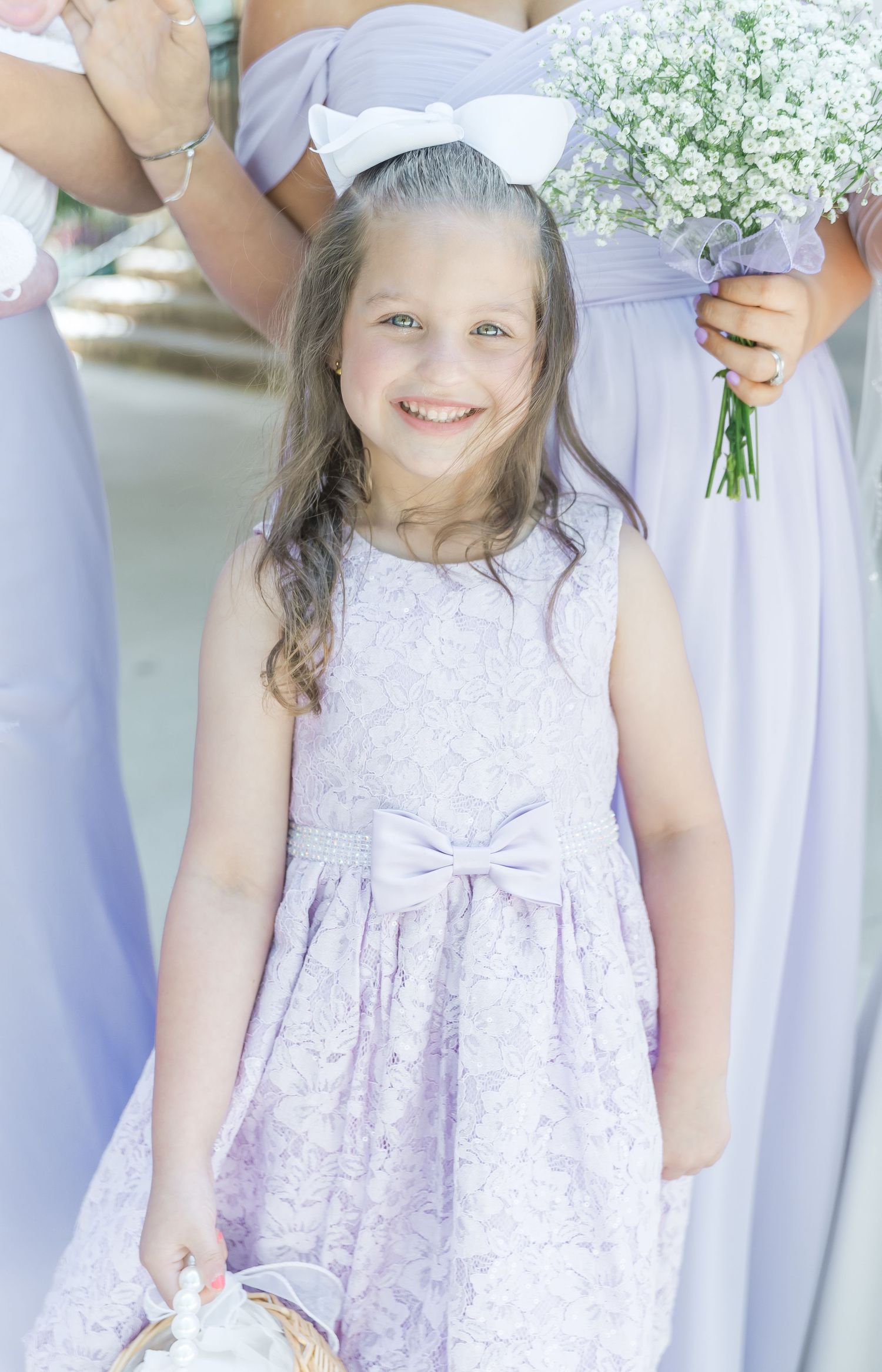 Young attendant in white lace dress with bow holds flower basket at outdoor wedding.