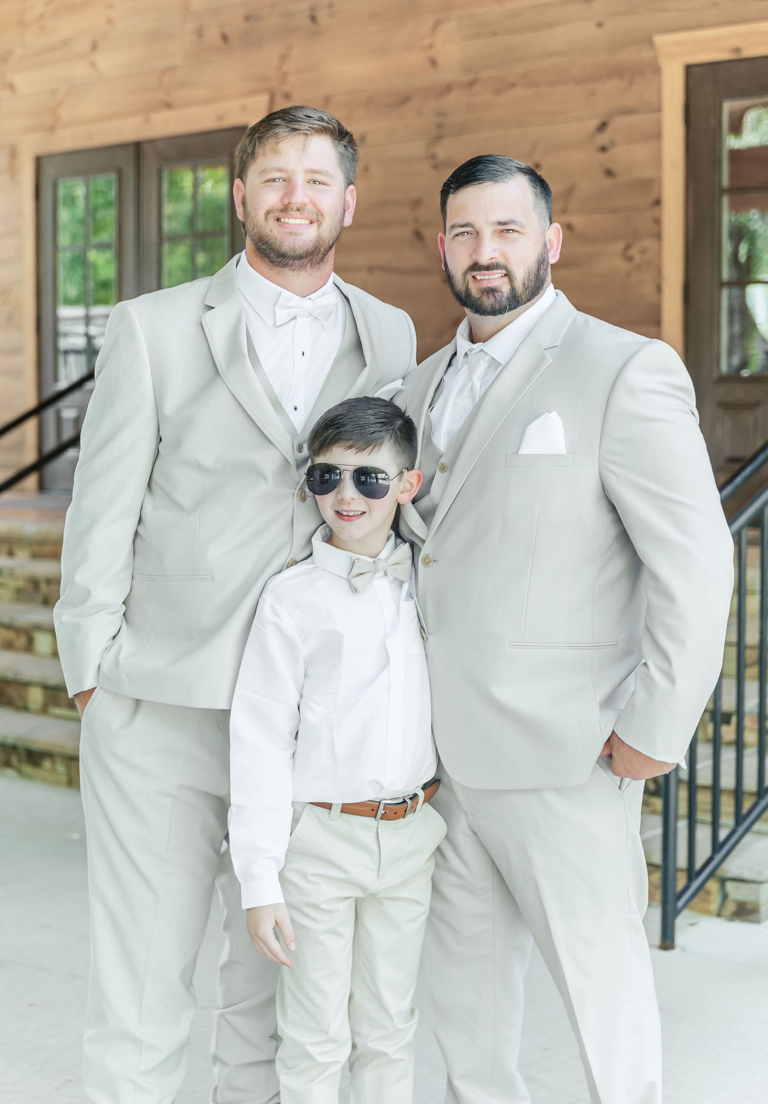 Three people in coordinating white and cream formal attire pose together outside.