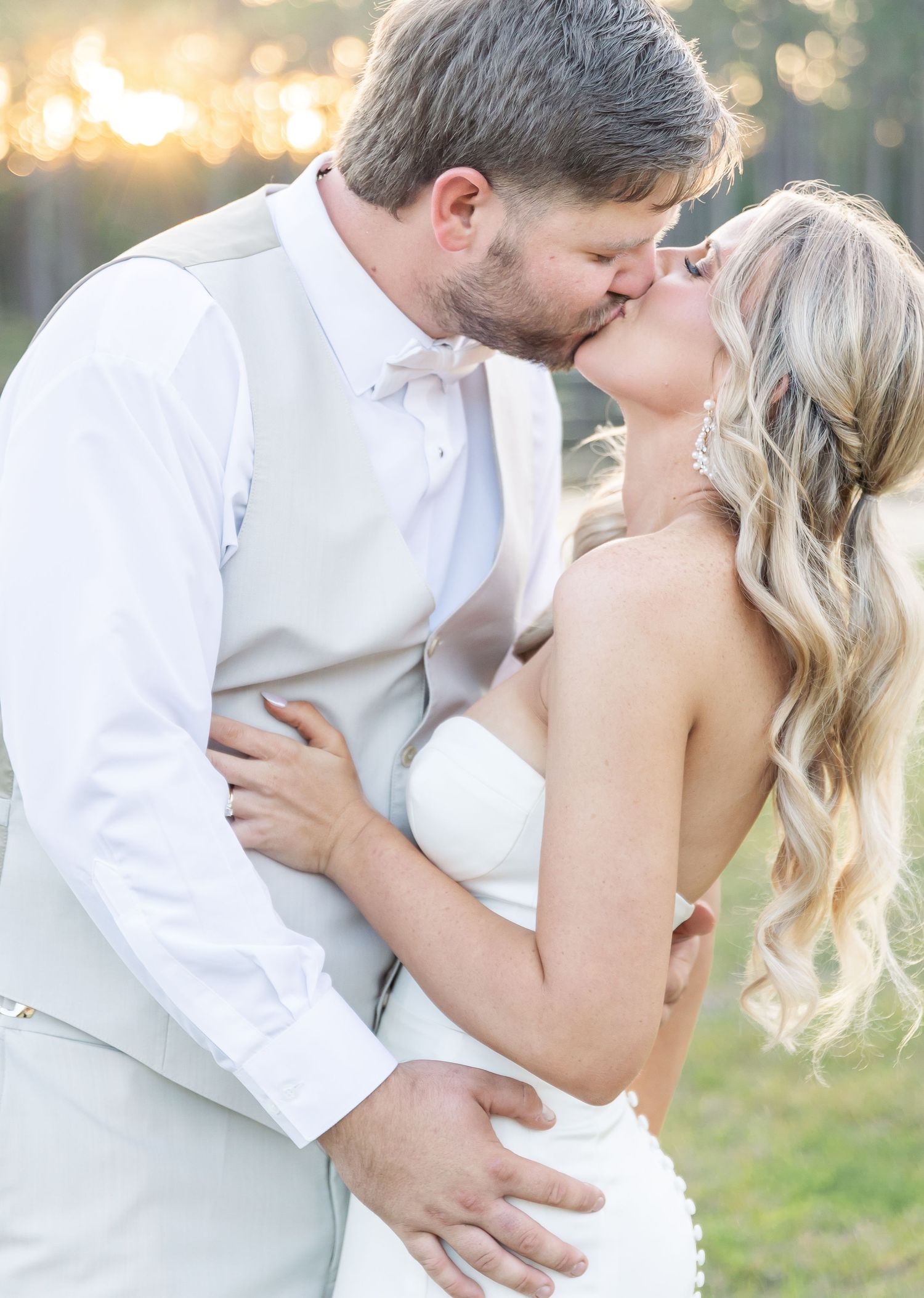 Intimate wedding portrait showing couple in white sharing a kiss with golden sunlight filtering through trees.