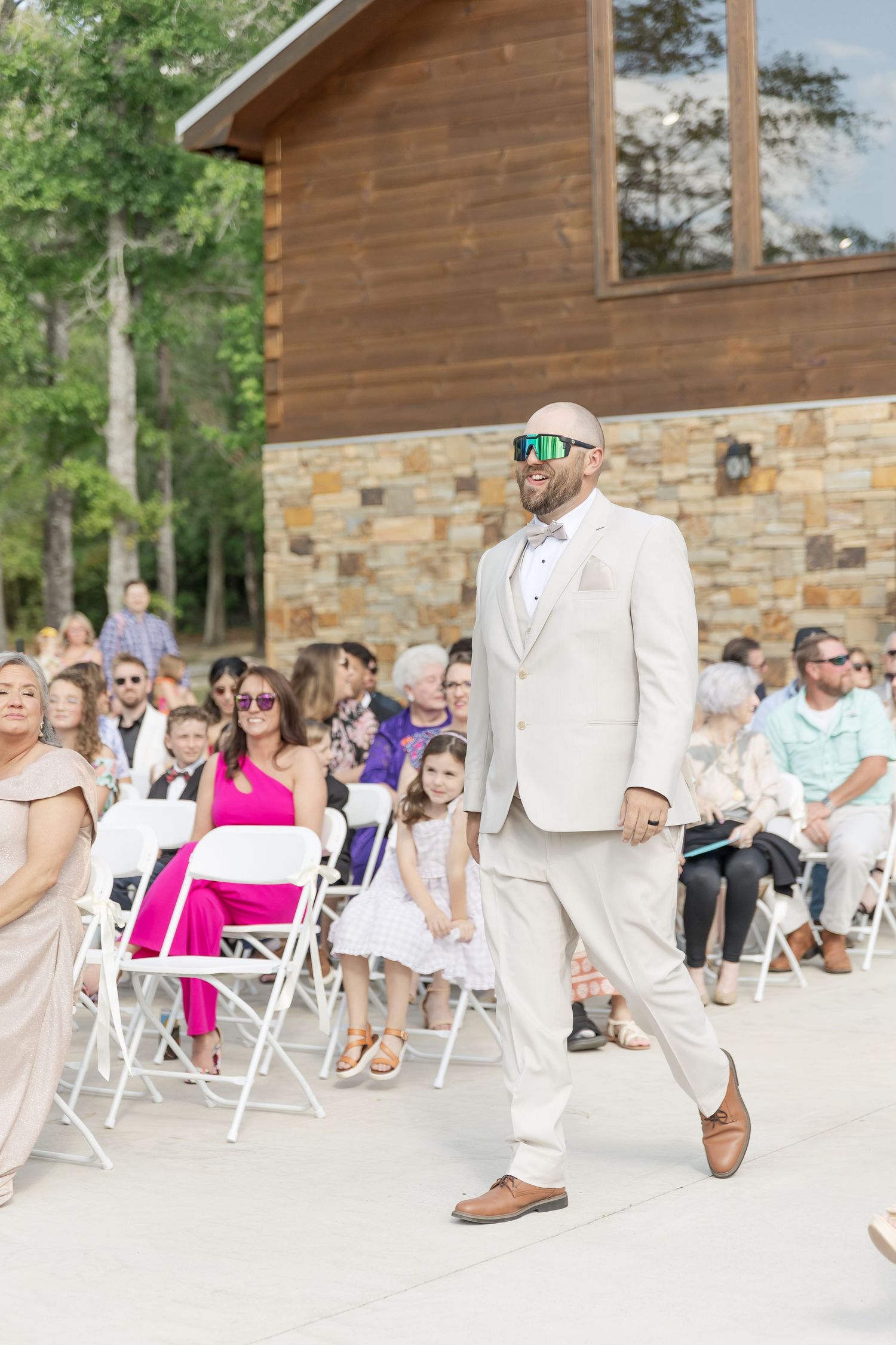 Wedding party member in cream suit and green sunglasses walks past guests in white folding chairs.