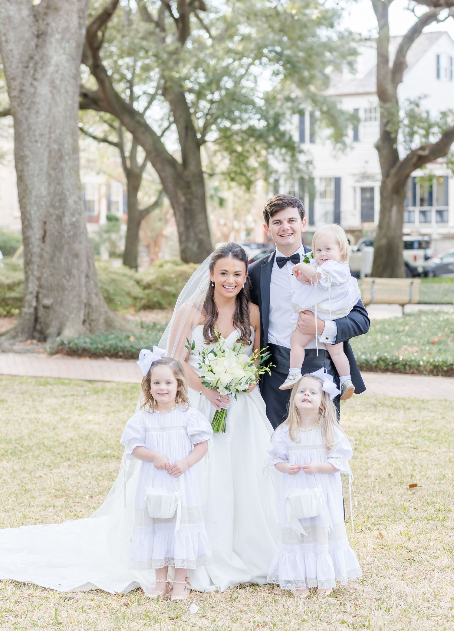 Bride and groom pose with three young children in flower girl dresses outdoors at a sunny wedding celebration.