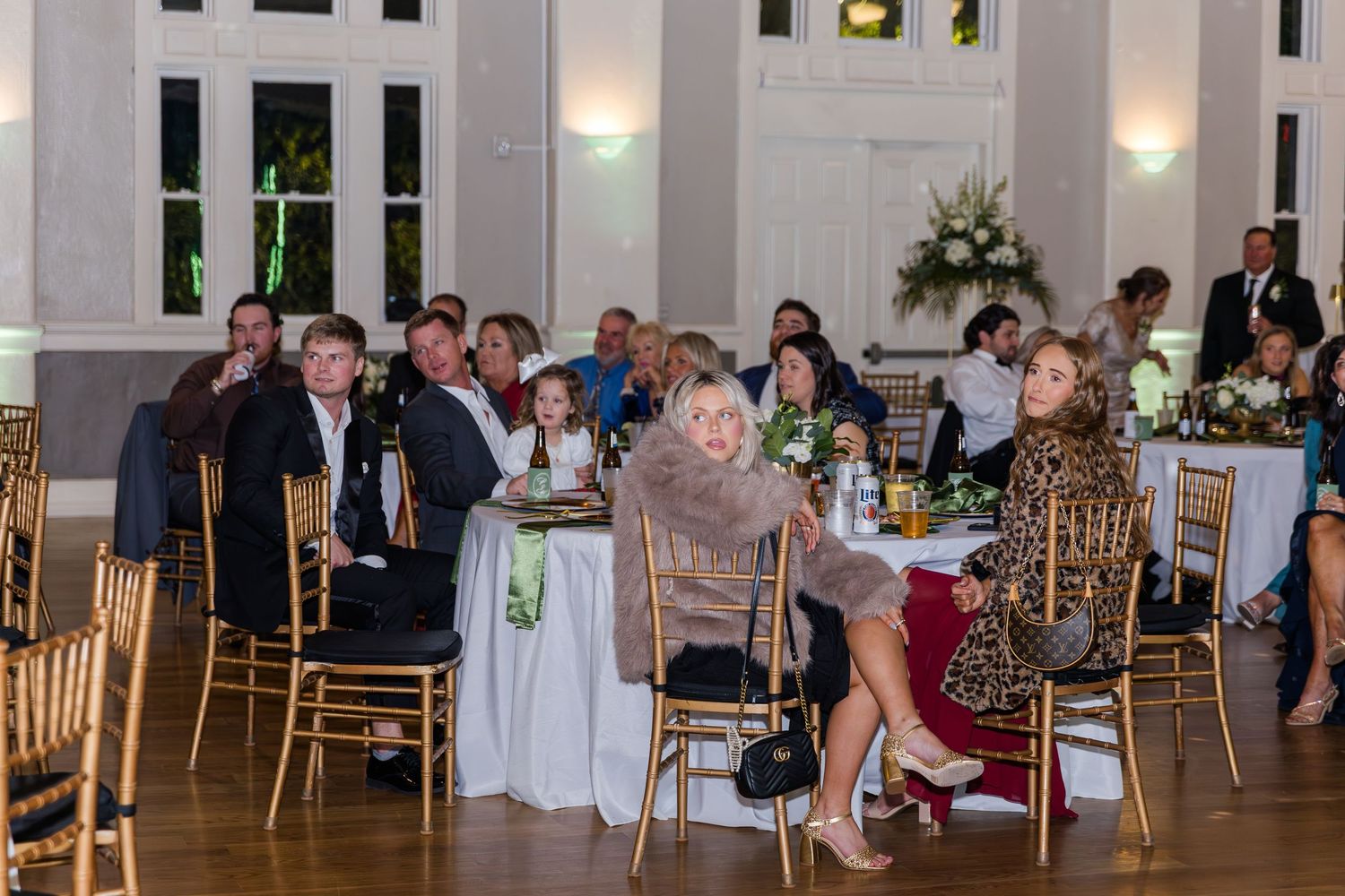 Guests seated at round tables during an elegant indoor wedding reception in a grand ballroom with gold chairs.