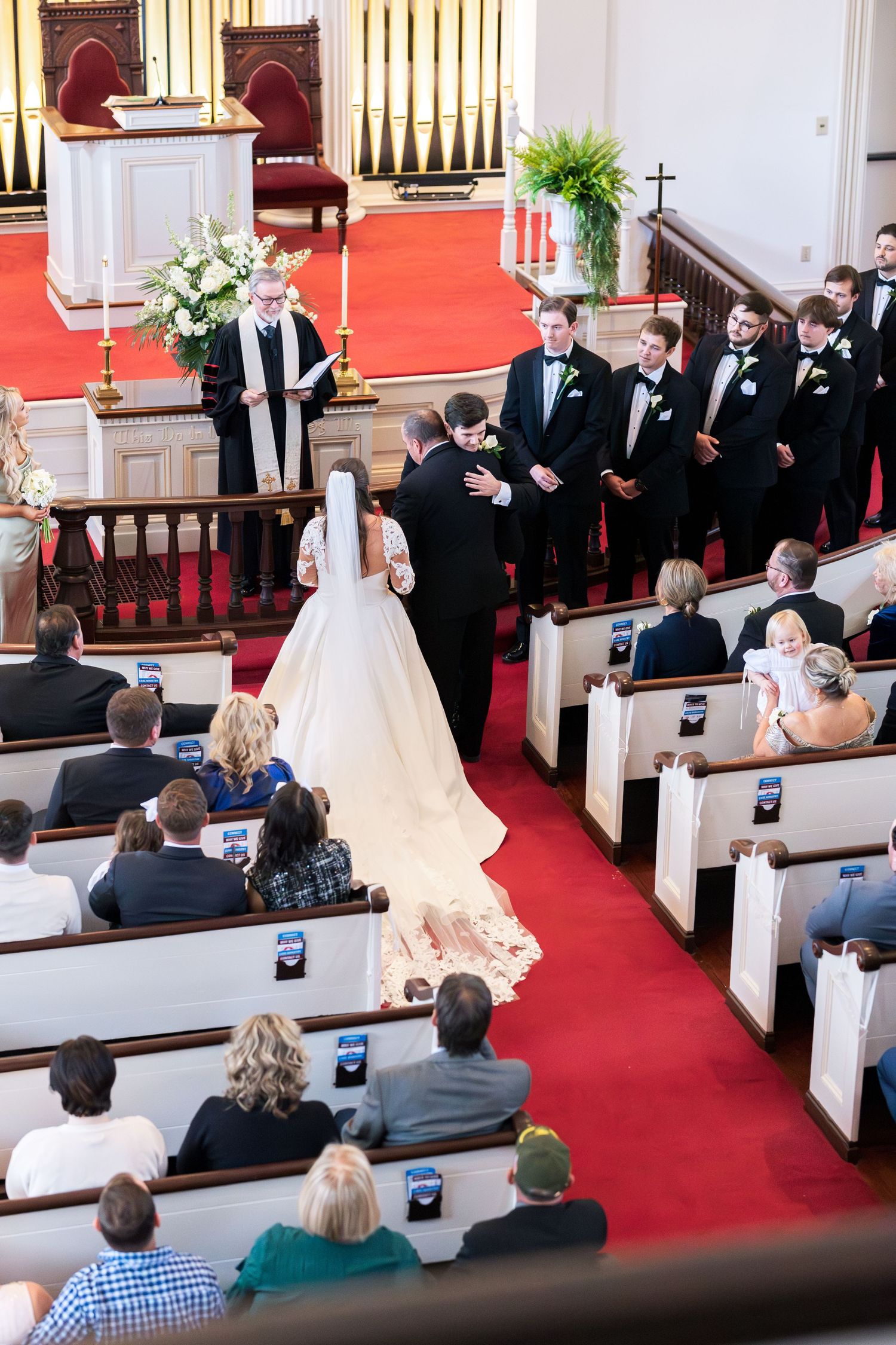 Bride in white gown walks down red carpet aisle toward groom at church wedding ceremony.