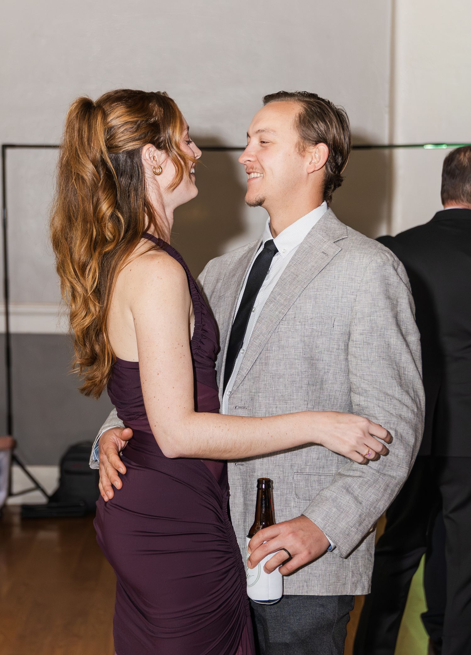 Couple slow dancing at wedding reception, man in gray suit holding beer bottle.