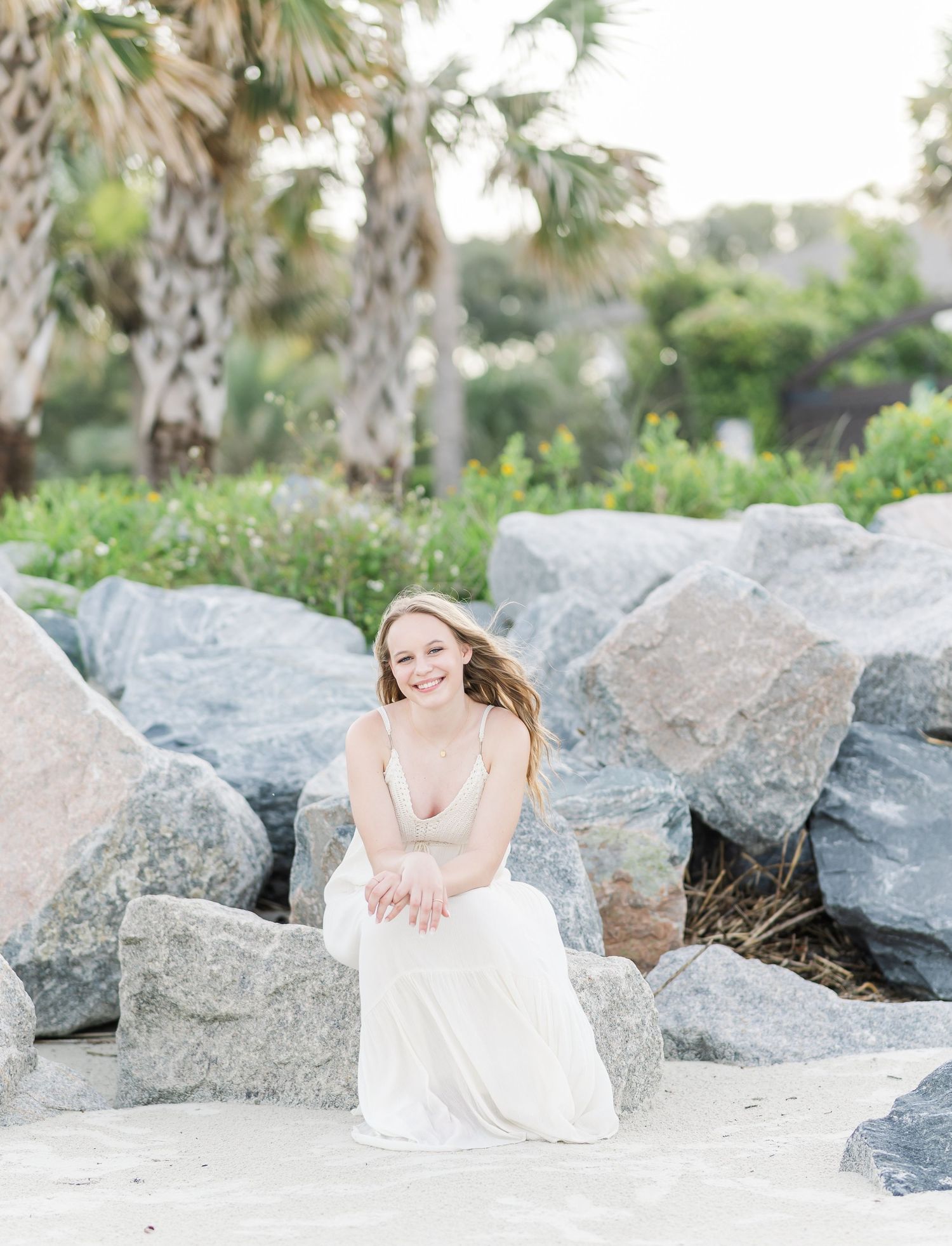 A serene coastal photoshoot set among large rocks and palm trees with ethereal white dress flowing in the breeze.