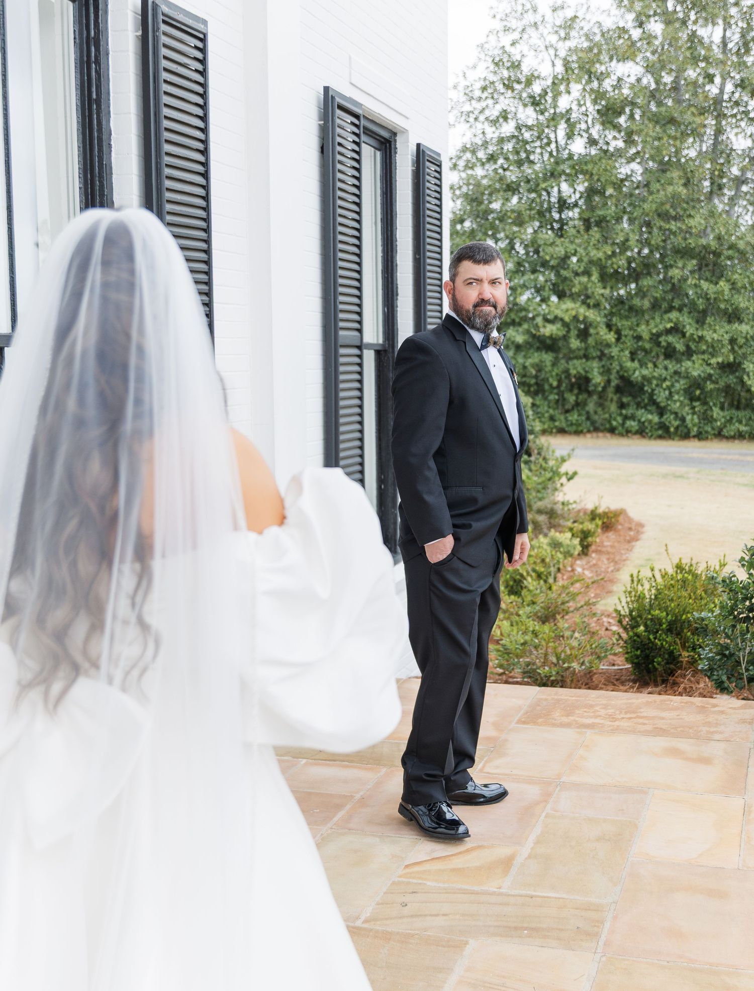 Groom in black tuxedo turns to see bride in white gown and veil during first look at outdoor wedding venue.
