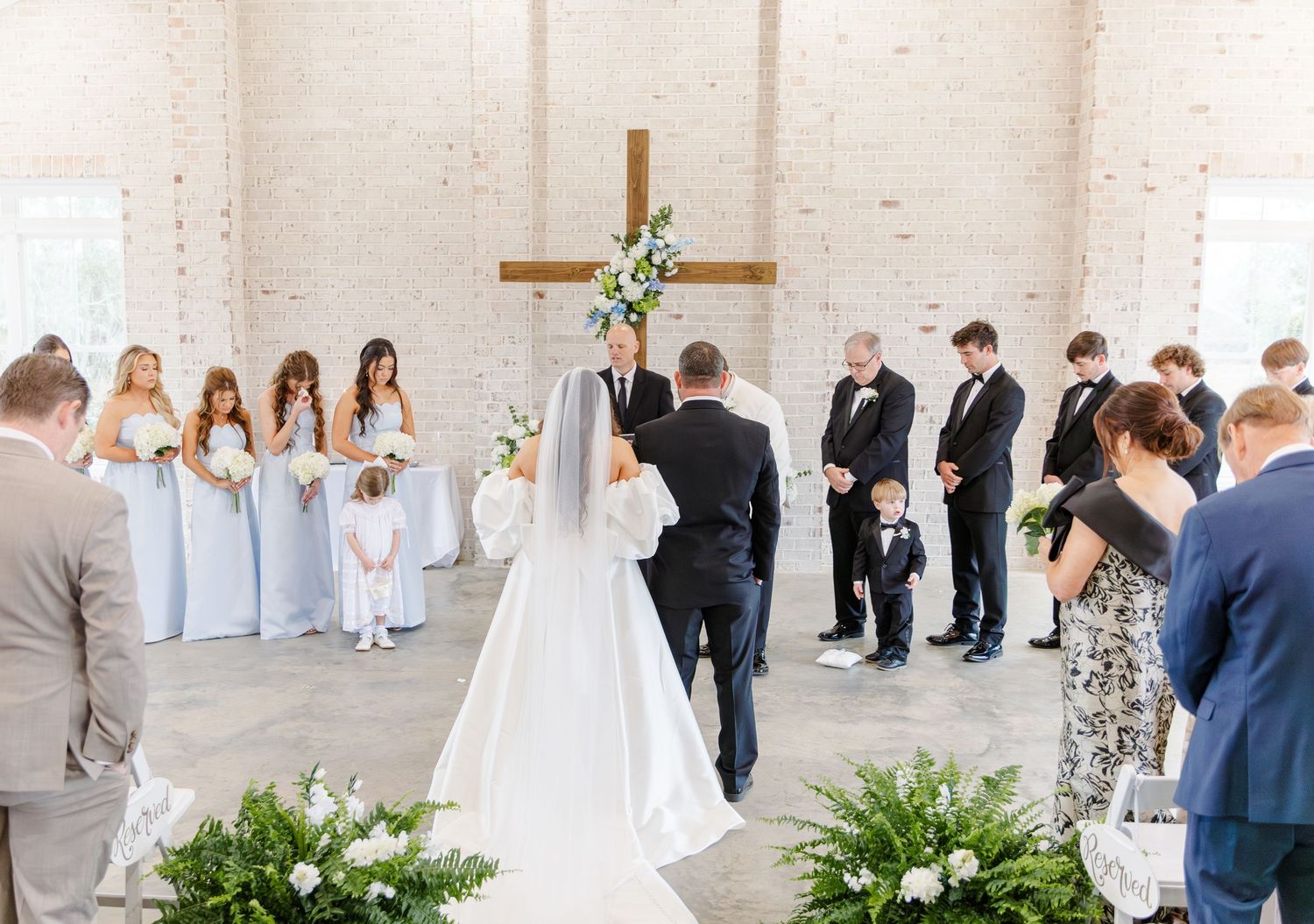Wedding ceremony with bride and groom at altar, bridesmaids in blue, groomsmen, and flower girl in white chapel.