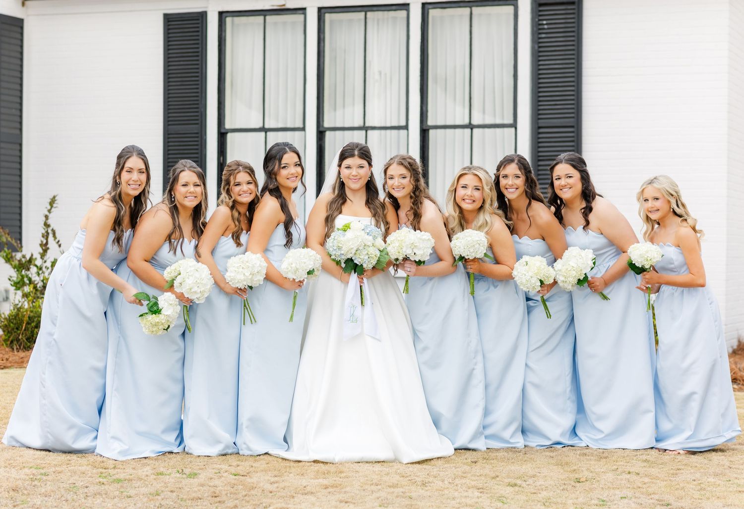 Bride poses with eight bridesmaids in matching powder blue strapless gowns holding white floral bouquets.